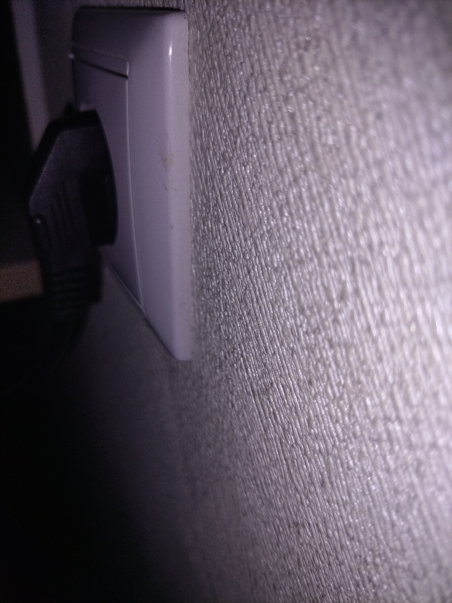 A close-up shot shows a white electrical outlet with a black plug inserted. The outlet is mounted on a textured wall. The lighting is dim, with the left side of the image mostly in shadow and the right side illuminated. The focus is on the outlet and the texture of the wall. The image is of a residential setting.