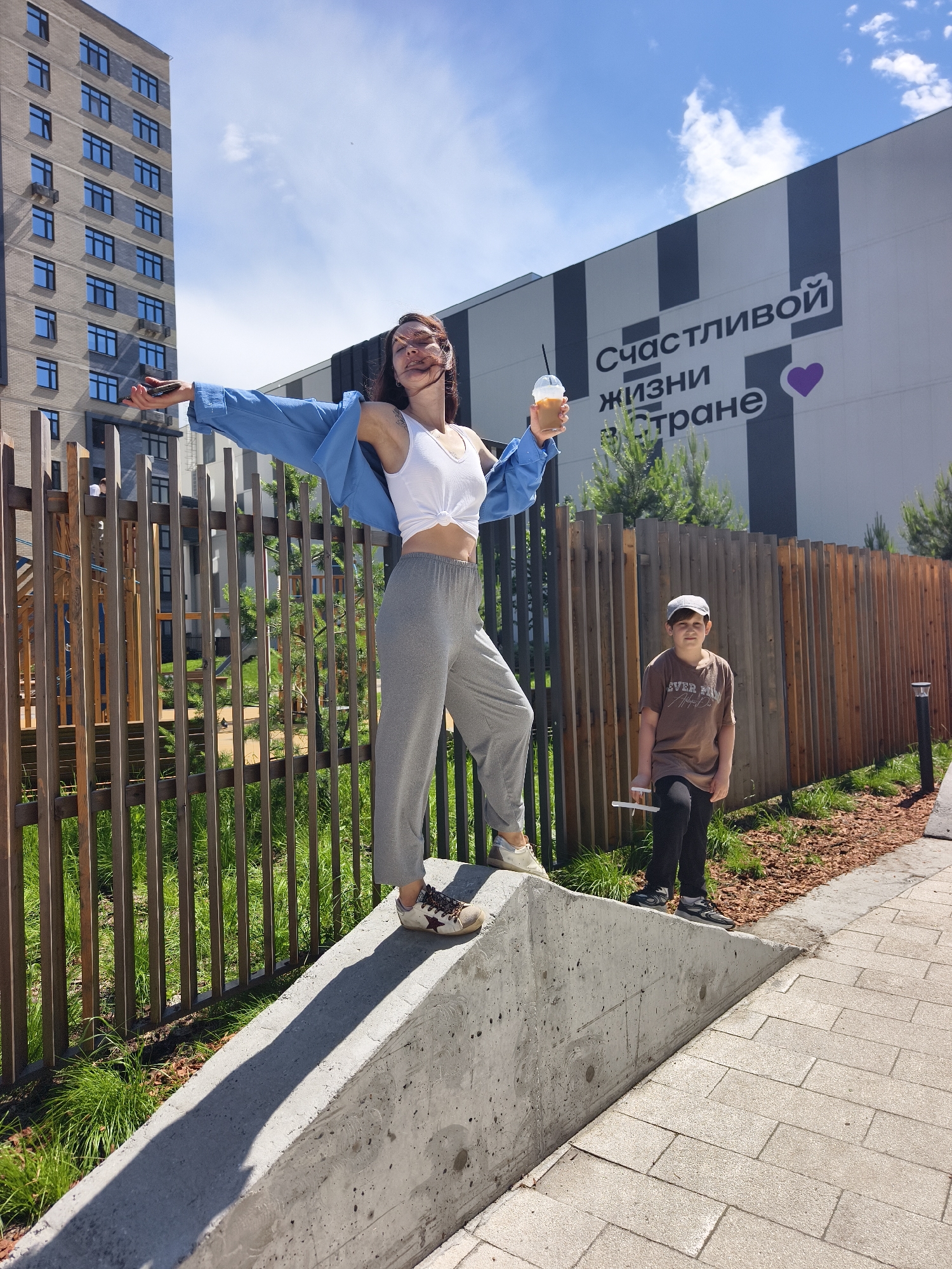 The image captures a sunny outdoor scene in Tyumen, Russia, featuring a young woman and a young boy in an urban setting.
The main subject, a young woman, stands dynamically on a concrete barrier with her arms outstretched. She has long, dark hair, wind-blown, and is dressed in a white knotted crop top, grey sweatpants, and a blue long-sleeved shirt worn open over her shoulders. On her feet are white sneakers with a distinct star symbol. She holds a clear cup containing an iced beverage with a straw in her right hand and a smartphone in her left. Her pose suggests a moment of joy or freedom.
To the right, a young boy stands on a paved walkway next to the concrete barrier. He wears a light-colored baseball cap, a brown t-shirt with "EVER FREE" printed on it, black pants, and dark sneakers. He holds a white stick-like object in his right hand and looks towards the camera with a neutral expression.
The background features modern architecture and landscaping. On the far left, a tall, multi-story residential apartment building with numerous windows rises. In the center-right background, a large commercial or public building with a black and white striped facade displays prominent Russian text: "СЧАСТЛИВОЙ ЖИЗНИ В СТРАНЕ" (HAPPY LIFE IN THE COUNTRY/LAND), accompanied by a purple heart symbol. A wooden slatted fence runs behind the subjects, separating a green, grassy area from the paved surfaces. Strong shadows cast by the concrete barrier indicate bright, sunny daytime weather with some clouds visible in the blue sky.