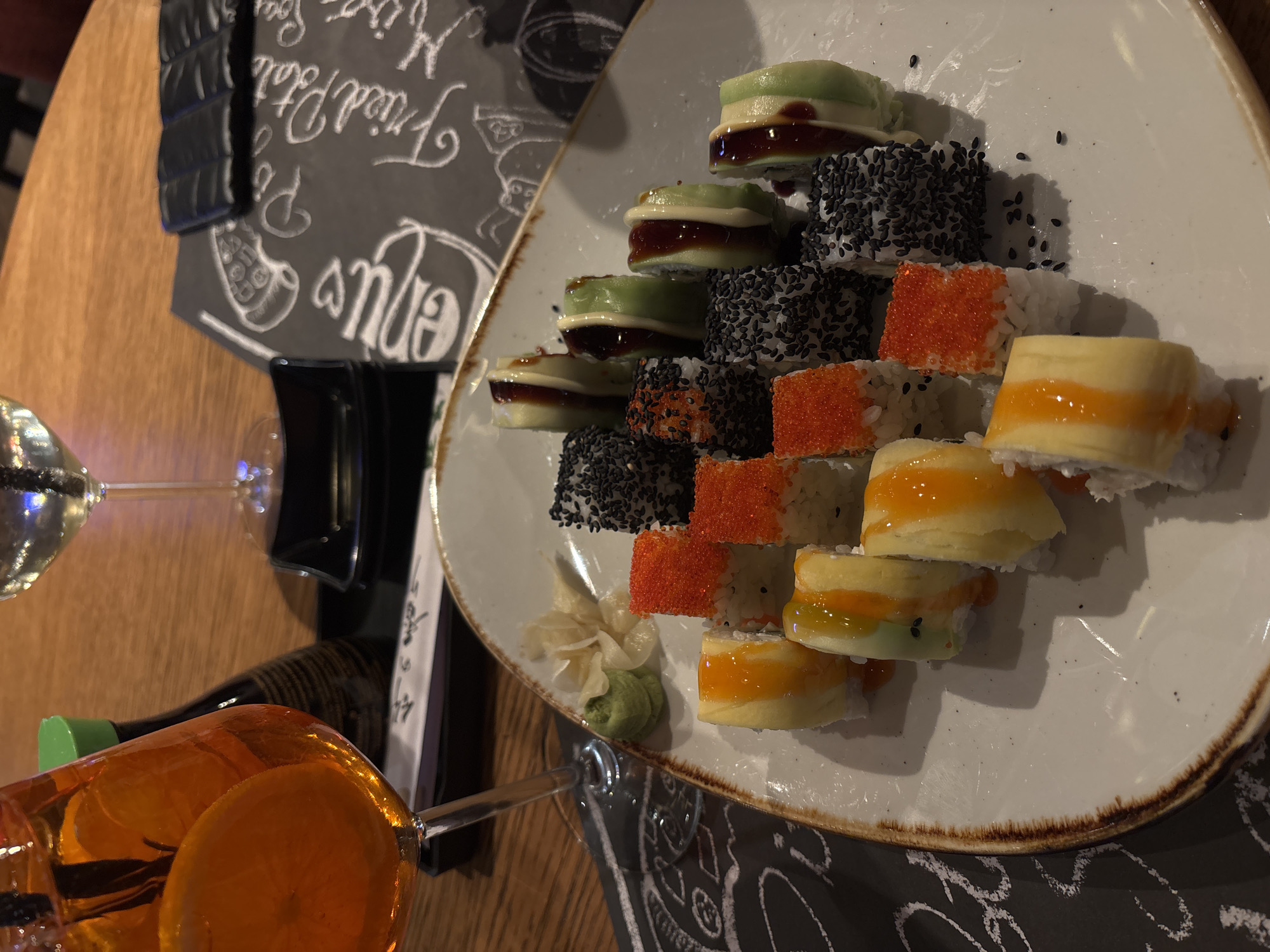 The image depicts a plate of sushi arranged in a pyramid shape on a white plate with a brown rim. The sushi consists of different varieties, including those with avocado, sesame seeds, and mango slices. Next to the sushi, there is a small portion of pickled ginger. A glass of orange-colored liquid with an orange slice is placed beside the plate. In the background, there is a menu card with white writing on a dark background. A glass of white wine is also visible. The setting suggests a restaurant or dining environment. Due to the angle and limited visual cues, it's challenging to definitively confirm the location as Varna, Bulgaria, without additional contextual information.