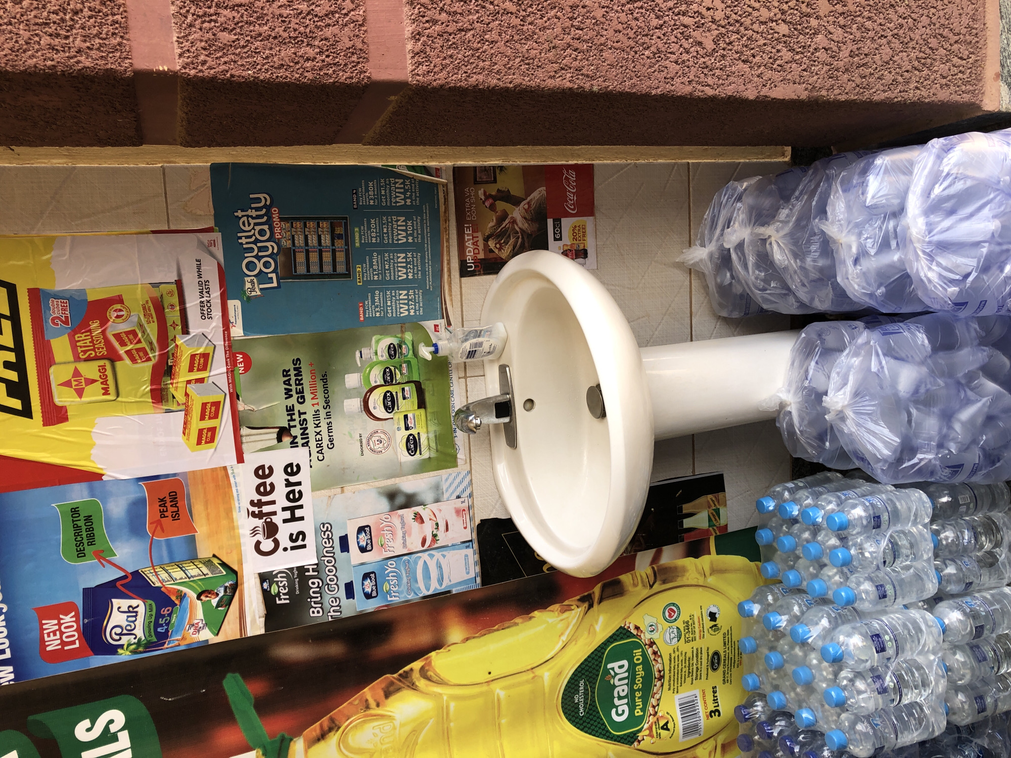 In the city of Bar Jirgi Summa, Nigeria, a brightly lit outdoor scene unfolds. A white pedestal sink with a chrome faucet is positioned against a wall adorned with various advertisements. To the left, a large yellow bottle of Grand Pure Soya Oil stands prominently, featuring nutritional information and certifications. To the right of the sink, stacks of bottled water, some in plastic bags, are visible.
The advertisements plastered on the wall showcase a range of products. One poster promotes Peak milk with a "New Look" and "Descriptor Ribbon." Another features Maggi Star Seasoning with an offer of "2 double cubes FREE." A colorful advertisement for "Outlet Loyalty" with a "PROMO" is also present, detailing reward bands for purchases. Smaller ads include "FreshYo Drinking Yogurt," and "Carex Kills 1 Million Germs in Seconds." An advertisement for Coca-Cola with a "20% EXTRA FREE" offer is also visible. The wall itself appears to be textured and reddish-brown. The ground beneath the sink and bottled water is paved, with some debris visible. The overall impression is one of a commercial or roadside stall in Nigeria.