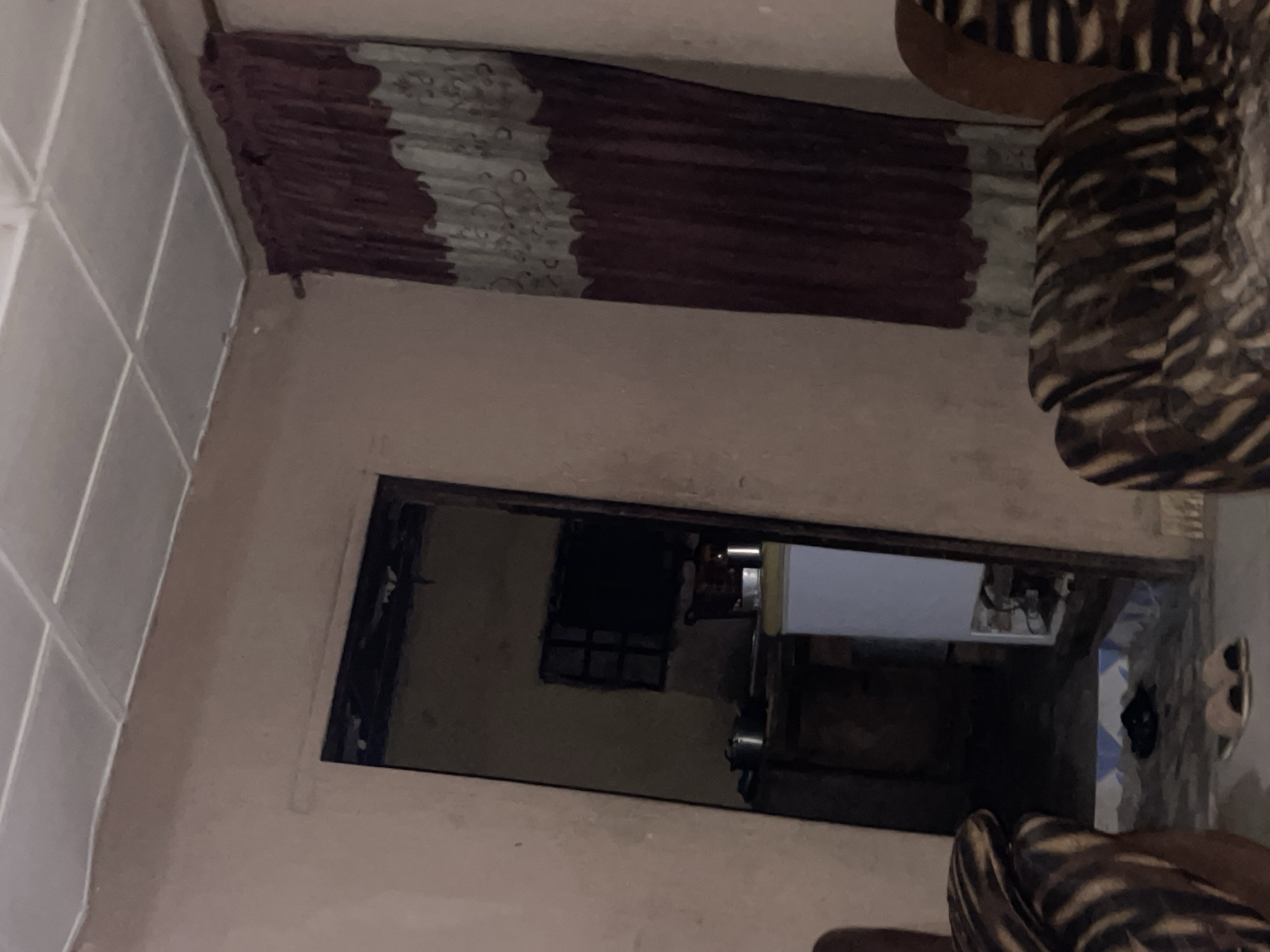 The image depicts an indoor scene, likely a modest residential interior in Lokoja, Nigeria. The room is dimly lit, with a textured, pale pink wall on the left and a large curtain with burgundy and grey patterned sections on the right. The curtain hangs from a rod. A doorway on the left reveals a glimpse into another room, which appears to be a kitchen or storage area, containing pots and possibly a refrigerator. The floor in the foreground has a tiled pattern. Two upholstered chairs with a dark brown and tan animalistic print are visible, one on the left and one on the right edge of the frame. A pair of light brown slippers lies on the floor in front of the doorway. The ceiling is paneled with white rectangular tiles. The overall impression is one of a simple, lived-in domestic space.