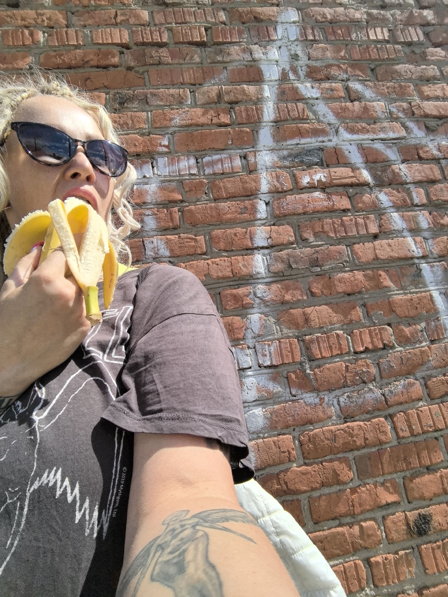 A woman with blonde braided hair, wearing sunglasses and a dark t-shirt, eats a banana in front of a brick wall. The wall is made of red bricks with white spray paint markings. The woman has a tattoo on her arm. The setting appears to be outdoors.