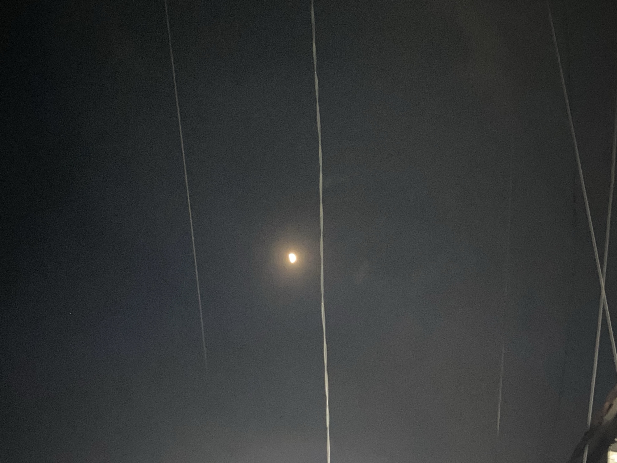The image shows a nighttime sky with a full moon visible near the center. The moon appears as a bright, slightly fuzzy disc. The sky is dark and overcast, with a hazy atmosphere. Several thin, white lines, possibly power lines or wires, run vertically through the frame, obscuring parts of the sky and the moon. One of these lines appears to be particularly close to the camera and is thicker and more detailed than the others, suggesting it is a foreground object. To the left of the moon, a faint, tiny white dot is visible, which could be a star or a distant light. The overall mood of the image is quiet and somewhat mysterious due to the darkness and the obscuring lines. The presence of the power lines suggests an urban or semi-urban setting. Given the location context of Madiun, Indonesia, this could be a view from a balcony, window, or street at night. No text is visible in the image.