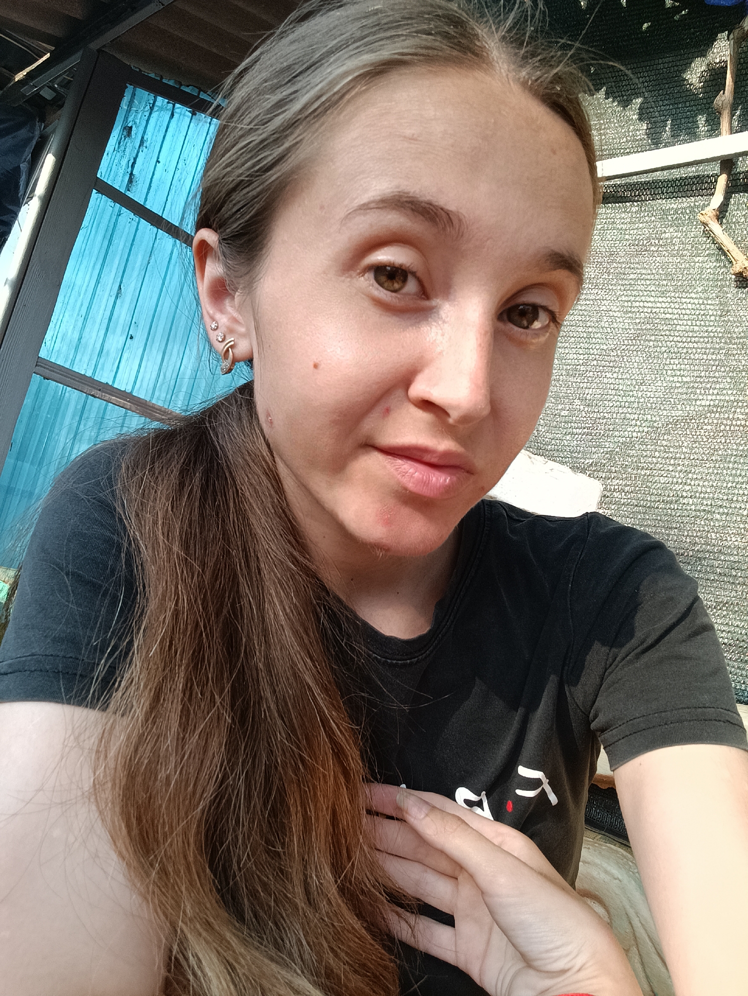 The image shows a close-up, selfie-style shot of a young woman with long, light brown hair tied in a ponytail, and brown eyes. She is wearing a black t-shirt. Her left ear is visible, adorned with multiple earrings. The background is out of focus but appears to be an outdoor setting with a blue, corrugated material on the left and a light-colored mesh material on the right, possibly indicating a shaded area or a fence. The lighting suggests it is daytime. The location is identified as Vypasne, Ukraine.