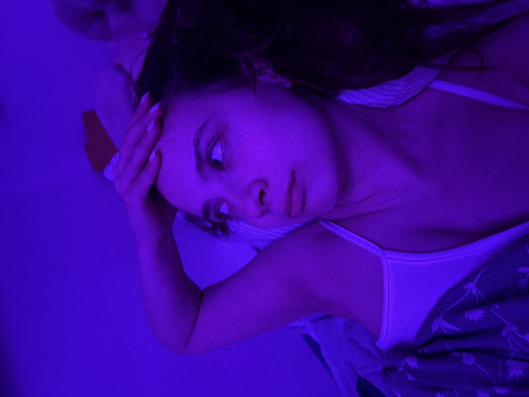 A woman lies in a bed, illuminated by blue light. Her dark hair is partially visible at the top of the frame. She wears a white tank top and a floral-patterned blanket covers part of her torso. Her right arm is raised, and her hand touches her forehead. Her eyes are directed to the left. The location is Прикамский, Russia.