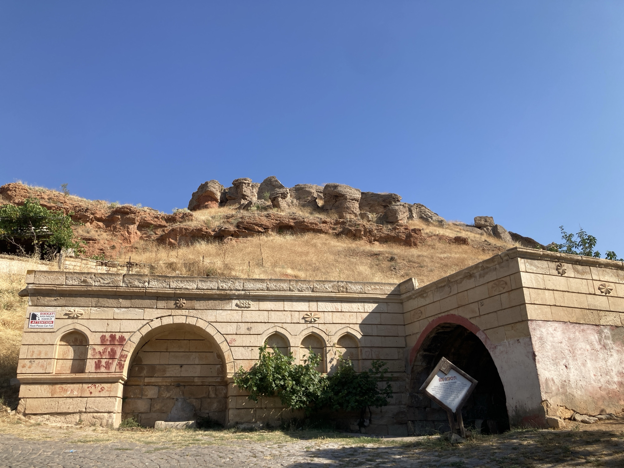 The image shows a historic stone structure in Avanos İlçe Merkezi, Turkey, under a clear blue sky. The structure features arched openings and is built with light-colored stone blocks. Above the arches, there is decorative stonework and a date, "1885," is inscribed. Red handprints are visible on the stone wall of one of the arched sections, and a sign with text, some of which appears to be in Turkish and English, is posted. A small green tree grows in front of the structure. In the background, a dry, grassy hillside with rocky outcrops rises towards the sky. The ground in front of the structure is paved with cobblestones.