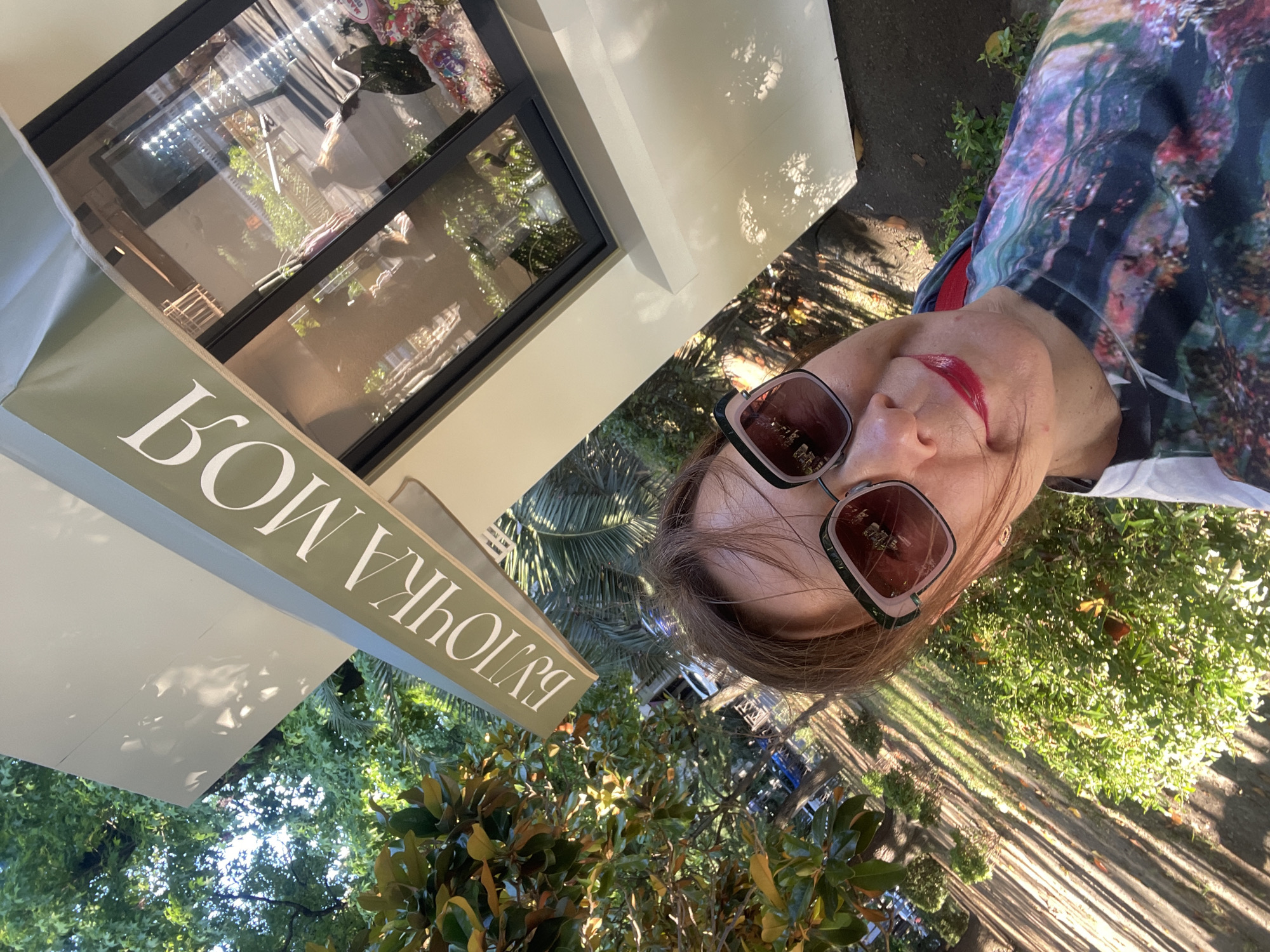 A woman with dark hair, wearing large sunglasses and red lipstick, takes a selfie outdoors. She is wearing a colorful, patterned t-shirt. Behind her, a small, modern kiosk with a green awning and large glass windows is visible. The awning has white Cyrillic text that reads "БУЛОЧКА МОЯ" (My Bun). The kiosk appears to be a bakery or cafe. The background shows lush green trees, palm fronds, and sunlight casting long shadows on a path. The setting is likely a park or street in Sochi, Russia, given the vegetation and architectural style.