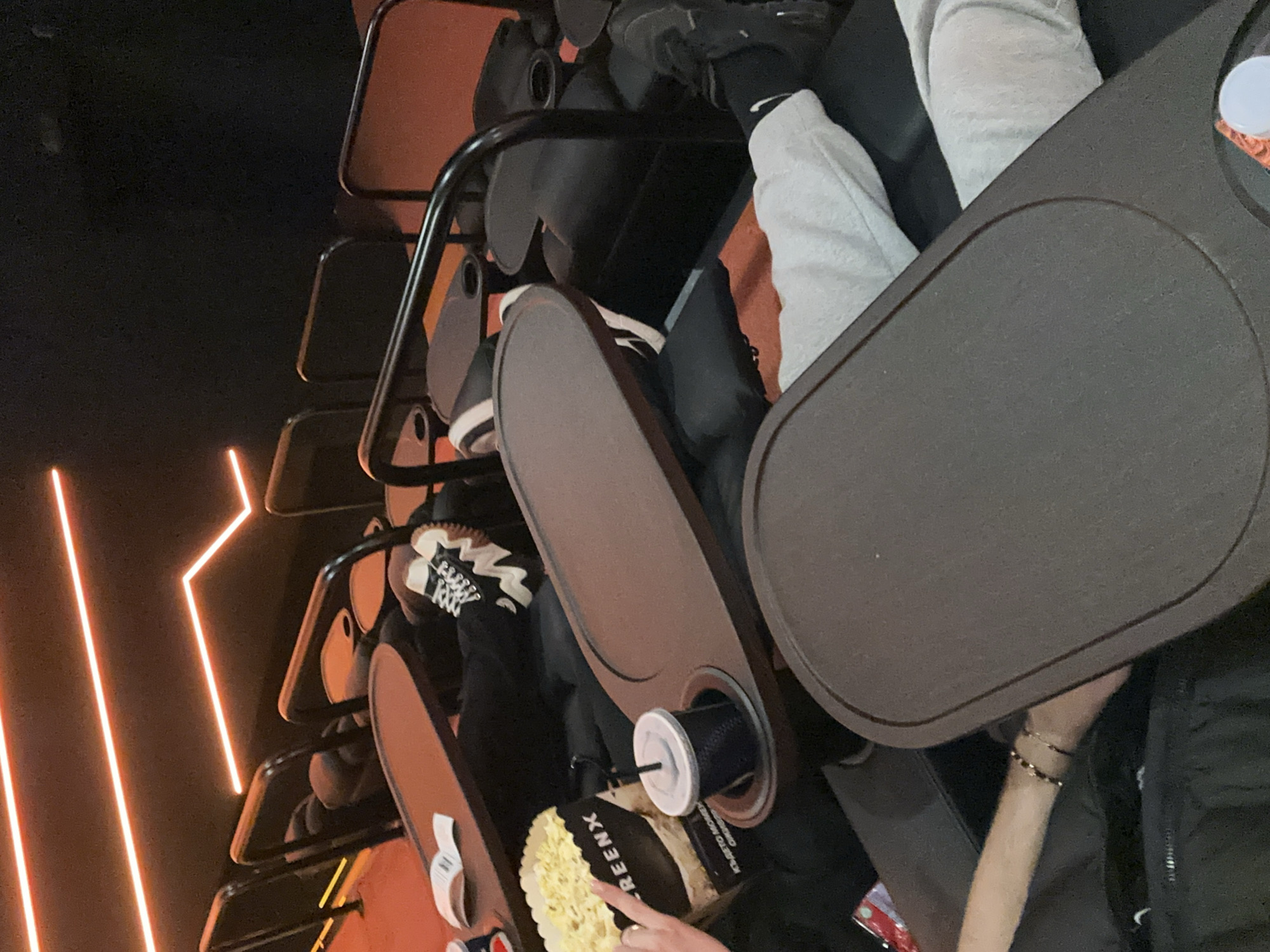 The image shows a view from inside a dark movie theater in Plovdiv, Bulgaria, likely in the Cinemax. The camera is positioned above the seats looking down at several rows. In the foreground, an individual is holding a bucket of popcorn and a soda with a straw in a cup holder. The theater seats have tray tables attached. A person is wearing grey sweatpants and black Nike socks, with black shoes visible further up. There are orange lights visible on the wall.
