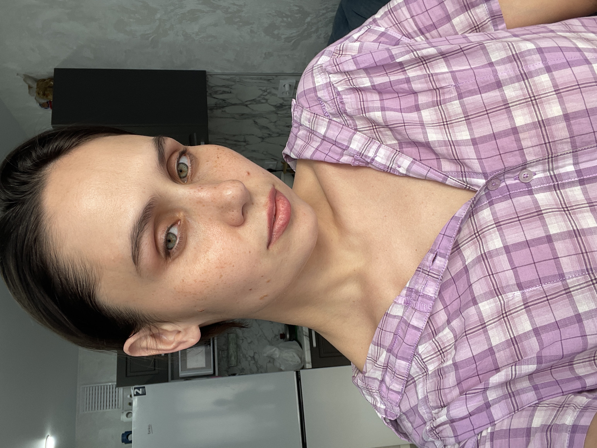 A young woman with short dark hair and freckles is the primary subject of the image. She is wearing a light purple and white plaid shirt. Her skin tone is fair. She is looking directly at the camera.
The background suggests an interior space. There is a black rectangular object mounted on the wall above a countertop, likely a kitchen setting, with marble-like tiles above the counter. Below the counter, a white refrigerator is visible. There is no evidence in the image to suggest the location is in Izhevsk, Russia.