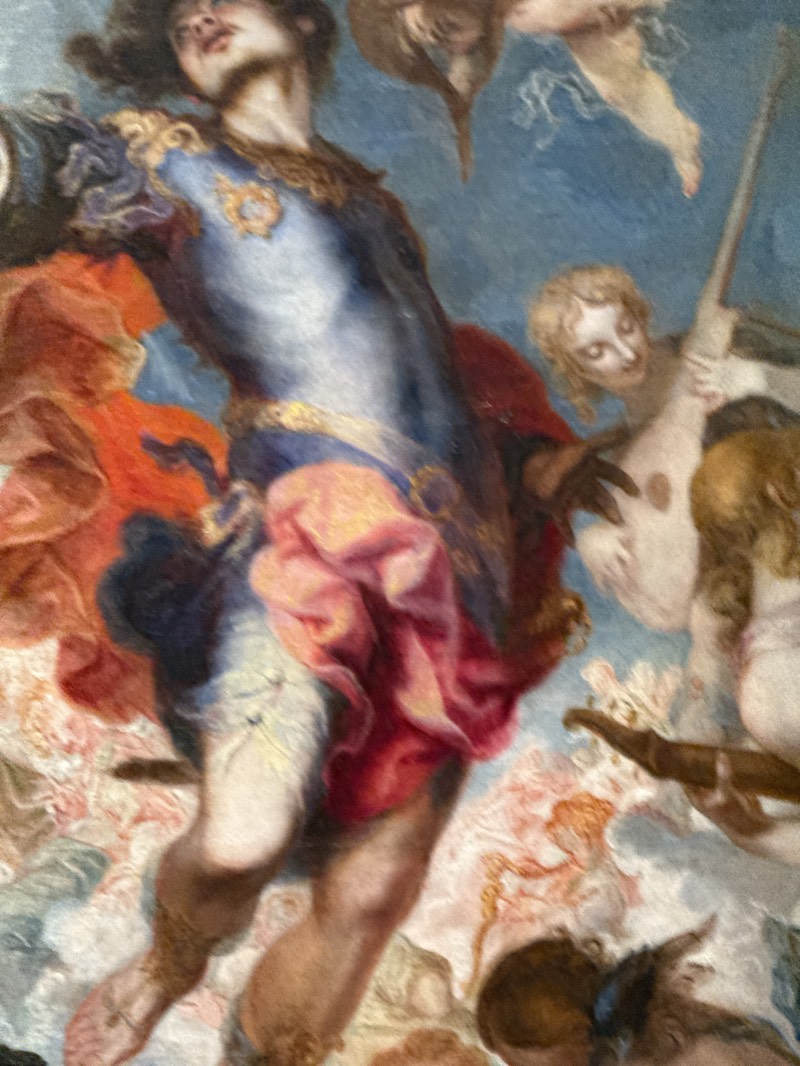 The image depicts a section of the ceiling fresco "The Apotheosis of Saint James" in the Church of San Antonio de los Alemanes in Madrid, Spain. The fresco features a central male figure, likely representing Saint James, wearing silver armor with gold accents, a blue cloak, and a pink cloth draped around him. He is surrounded by numerous figures of angels and putti, painted in light and airy colors. One angel is playing a musical instrument. The background consists of a light blue sky with clouds. The overall scene evokes a sense of ascension and religious exaltation.