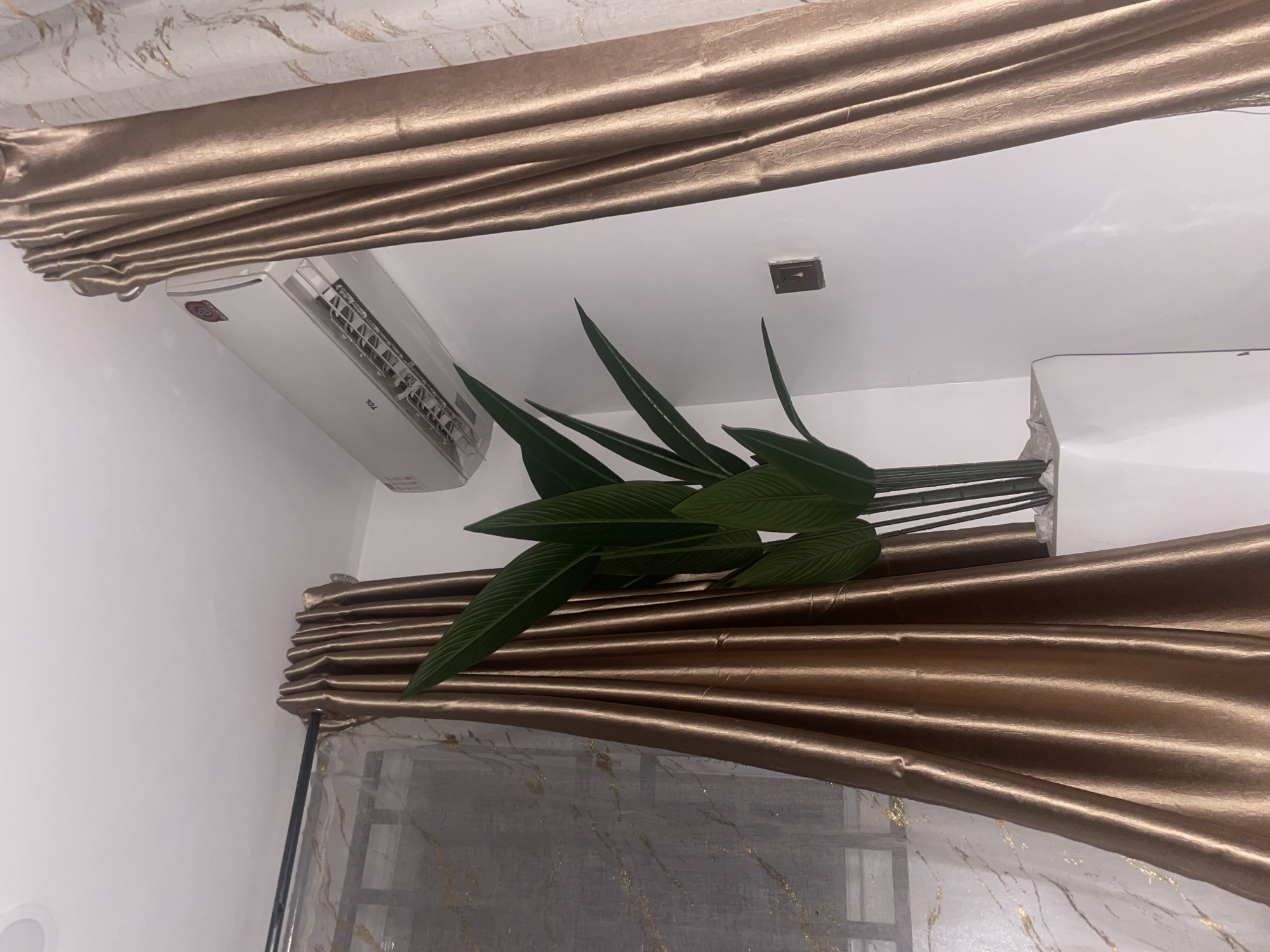 A low-angle shot reveals a room interior, likely in Kubwa, Nigeria, given the prompt. A white air conditioning unit is mounted on the wall. Artificial tropical leaves are positioned across the frame, partially obscuring the view. Brown curtains with a shimmering sheen hang from the ceiling and partially cover a window with a semi-transparent curtain. A ceiling fixture is also visible.
