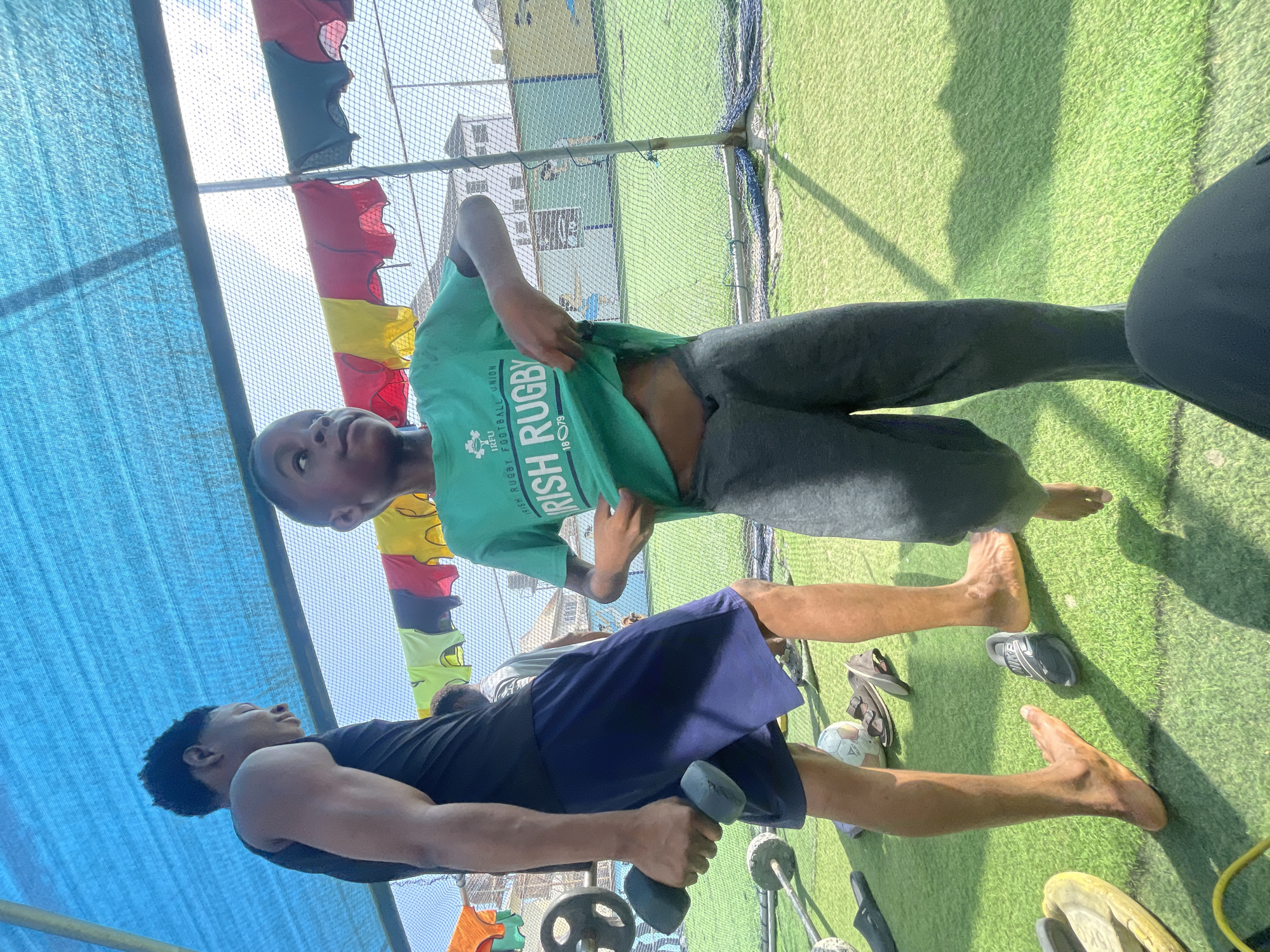 The image captures two young males engaged in physical activity at an outdoor training area in Iranla, Nigeria, under bright daylight. The ground is covered with artificial green turf. Overhead, a blue tarp or netting provides some shade, and in the background, a chain-link fence separates the area from what appears to be a building, with clothes (red, yellow, black) hanging on a line or fence.
In the foreground, a young male wearing a black sleeveless top and blue shorts is performing an exercise, holding a dumbbell in his right hand. He is barefoot, and his focus appears to be on his workout. Nearby, various weights, discs, and a soccer ball are visible on the turf, along with a pair of slippers and other shoes.
Standing slightly behind and to the left of the exercising individual, another young male wears a green t-shirt and dark grey sweatpants. He is also barefoot and is looking upwards, pulling at his t-shirt with his hands. The text "IRISH RUGBY" and "IRFU" is clearly visible on his green shirt. The strong sunlight casts distinct shadows on the turf. The overall scene suggests a community or personal fitness session in a warm, outdoor environment.