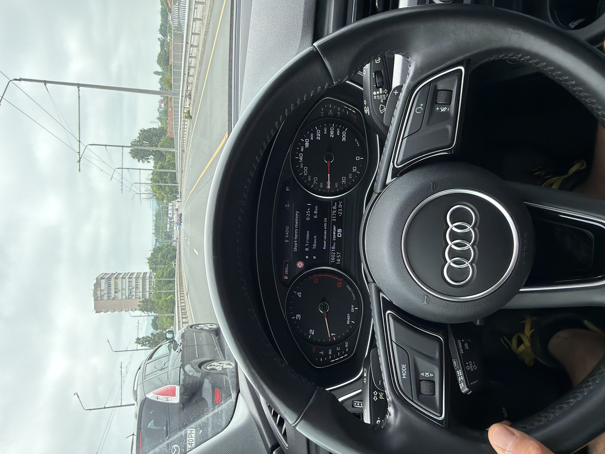 The image captures an interior view from an Audi car, showing the steering wheel and dashboard, with a partial view of the road and surrounding urban landscape in Burgas, Bulgaria.
A person's hand is visible on the lower right of the steering wheel, indicating an occupant, likely the driver or front passenger. The primary objects include the black leather-wrapped steering wheel with the Audi rings logo, the digital instrument cluster, and various control buttons and stalks.
The car appears to be stationary or moving very slowly, as the speedometer needle points to 0 km/h, and the tachometer shows the engine idling at approximately 1000 RPM. The instrument cluster displays "READY" and "D5" (drive gear). The short-term memory indicates a recent average speed of "16 km/h" over "0:25h" and "6.8km" distance, with a fuel consumption of "8.1 l/100km". The odometer reads "160218km", the trip meter shows "3179.8km", and the time is "14:57". The ambient temperature is "+23.0°C", and the estimated range is "360km".
Outside the car, a multi-lane road, possibly a bridge or overpass, is visible under an overcast sky, suggesting daytime. Power lines stretch across the view, indicating proximity to electric transport infrastructure. In the background, a tall, older residential building stands among trees, and the rear of another black car with a Bulgarian license plate "40 PH" (Plovdiv region code) is partially visible.