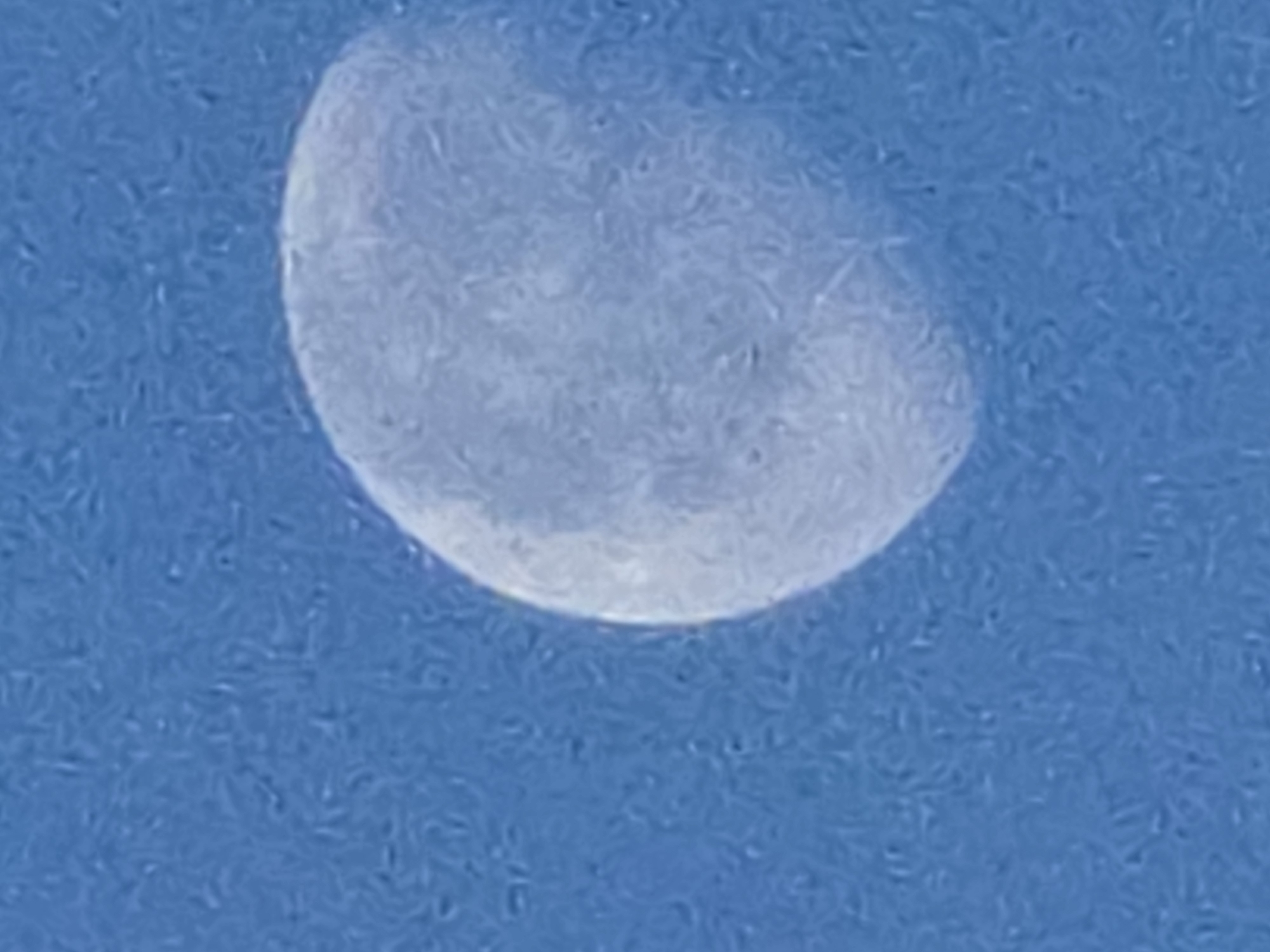The image shows a close-up view of the moon against a blue sky. The moon is partially illuminated, with a crescent shape visible. The surface of the moon appears textured, with visible craters and markings. The sky is a uniform blue, with no clouds or other celestial bodies visible. The image quality is somewhat grainy, suggesting it was taken with a low-resolution camera or under low-light conditions. There is no information provided within the image to suggest its location in Kelsterbach, Germany.