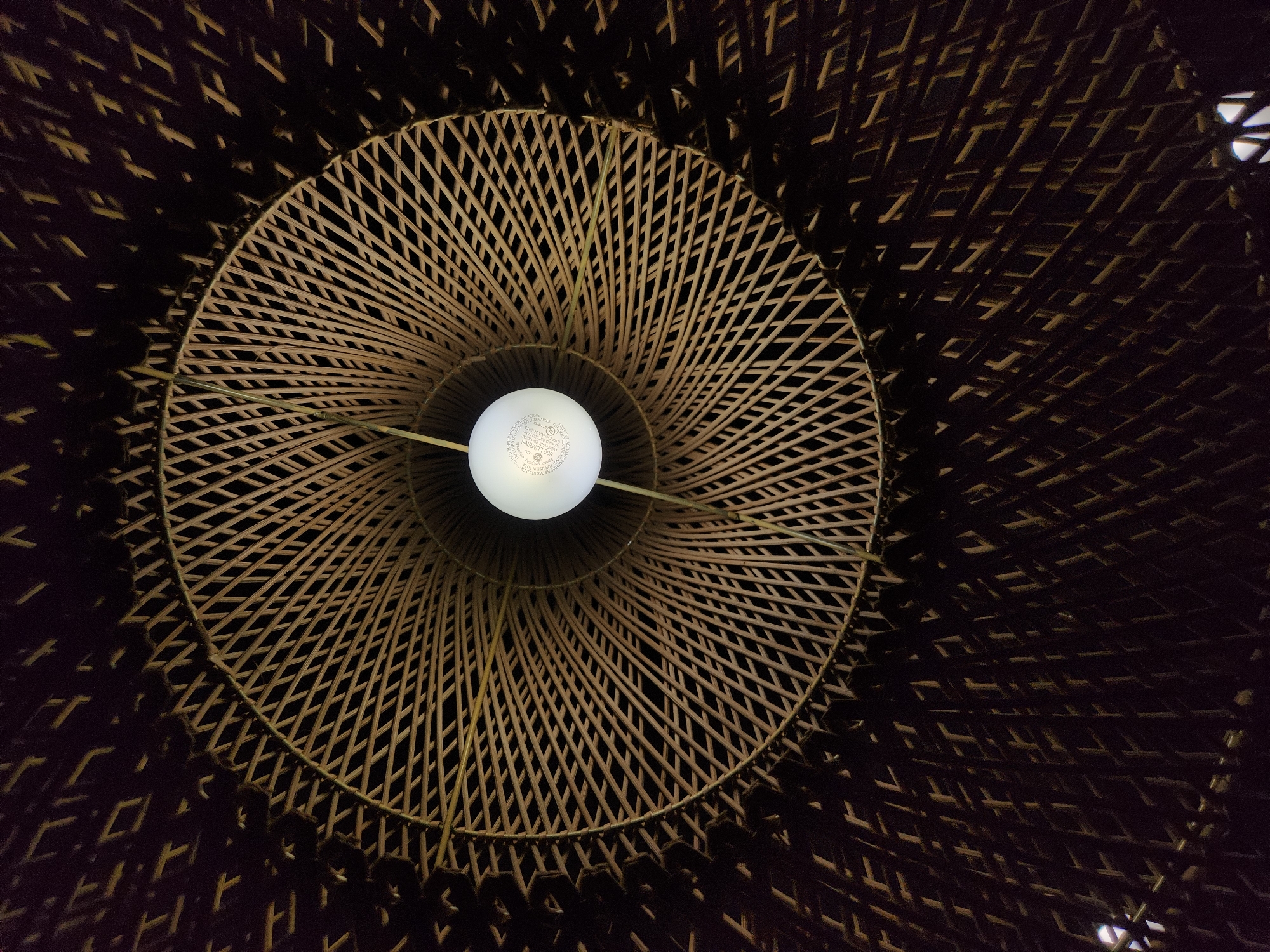 This is a close-up, low-angle shot looking up into a woven bamboo lampshade. A single, round LED light bulb is suspended in the center, emitting a soft, white glow. The lampshade has a complex woven pattern, creating a play of light and shadow. The bamboo strips are light brown and arranged in a radial pattern that fans out from the center. Towards the outer edges of the shade, the weaving becomes denser and darker, with larger openings that reveal glimpses of the dark background. The overall impression is one of natural materials and warm, diffused light. There is no visible text in the image. The scene appears to be indoors, likely a room illuminated by this fixture. The time of day and weather are not discernible.