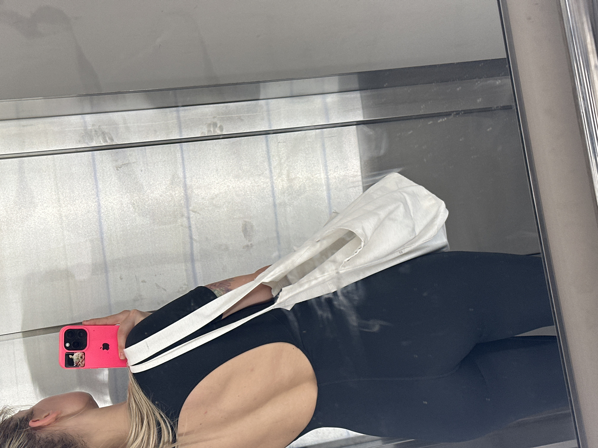 The image displays a mirror selfie of a person, likely female, captured indoors. The subject is seen in reflection, positioned horizontally, facing away from the viewer but looking towards her phone. She has blonde hair and is wearing a black, form-fitting garment, possibly a jumpsuit or athletic wear, which features a distinctive cut-out design revealing part of her lower back. A tattoo is visible on her upper right arm.
In her right hand, she holds a bright pink iPhone with a triple-lens camera array, angled to take the picture. The iconic Apple logo is visible on the back of the phone case, along with a small sticker or picture that appears to be an animal, possibly a dog. A white fabric bag, resembling a tote, with white straps, is slung over her left shoulder. Faint markings of the number "11" are discernible in several places on the white bag.
The background reflected in the mirror shows a plain, light grey or off-white wall above a horizontal metallic structure, which could be a shelf, beam, or part of a larger frame. The surface of the mirror is somewhat dirty, with visible smudges, streaks, and dust marks. Vertical lines are also present on the mirrored surface, possibly indicating panel divisions or minor imperfections. The scene depicts a casual moment, characteristic of a dressing room, gym, or private living space in Budva, Montenegro.