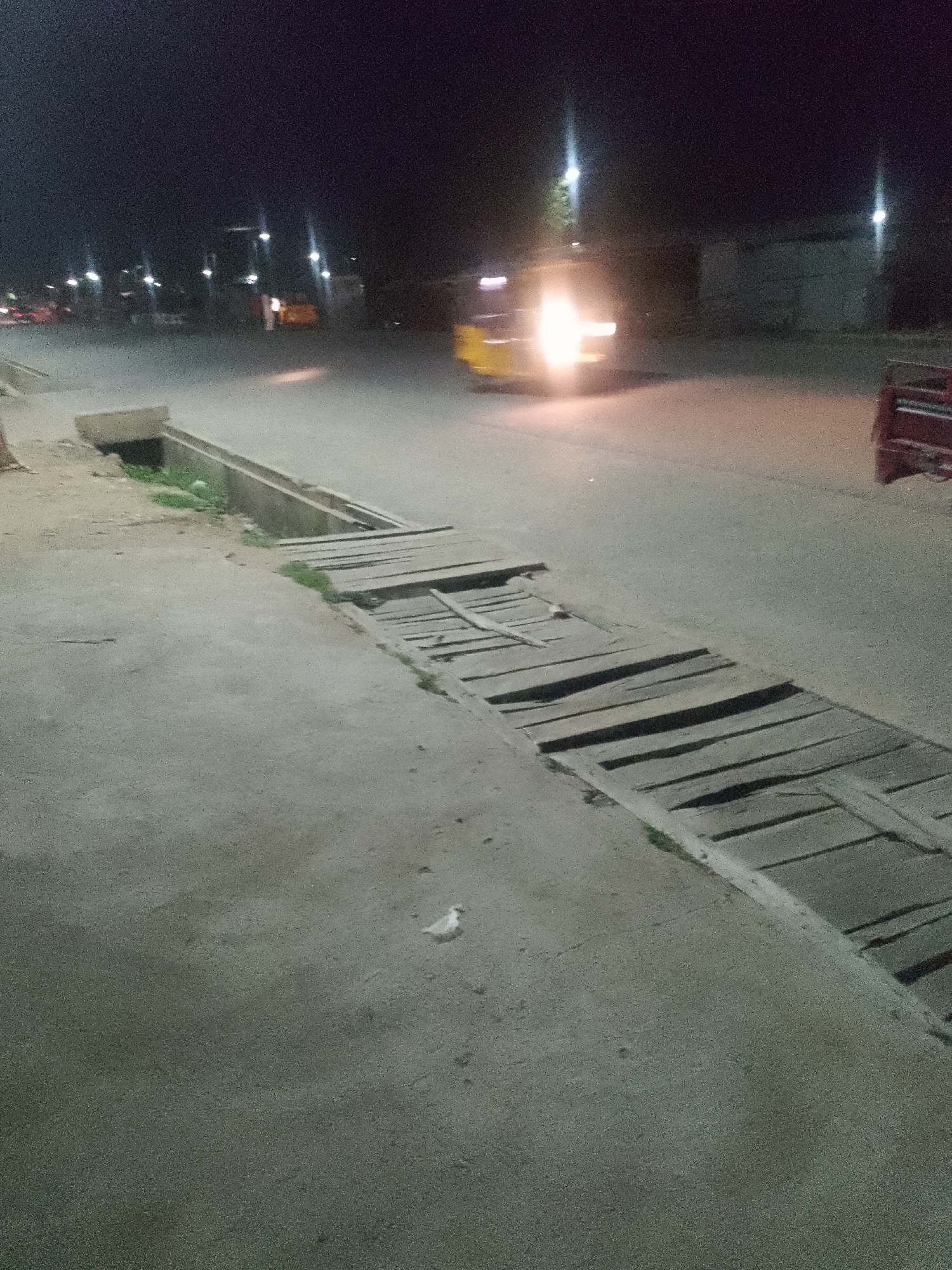 At night on a paved street in Jalingo, Nigeria, a yellow auto-rickshaw is moving, its headlights illuminating the scene. The street has a drainage channel with wooden planks covering a section of it. The surrounding area is dimly lit, with streetlights visible in the background. A red vehicle is parked on the right side of the image. The ground is a mix of concrete and rougher pavement. There is a small bird on the ground in the lower half of the image. The sky is dark.