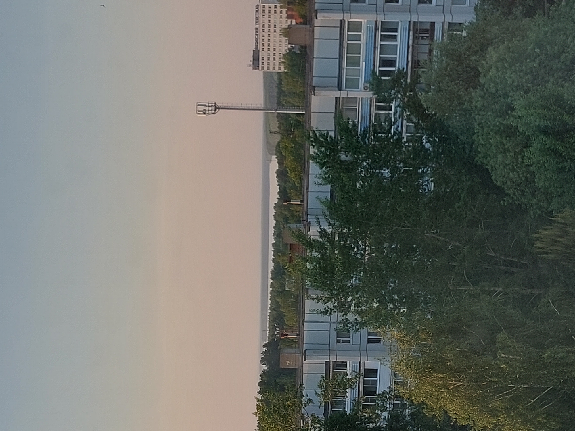 The image shows a view in Chaikovsky, Russia. The cityscape includes residential buildings with many windows, interspersed with green trees. There's a tall structure resembling a sports field lighting pole in the middle ground, and in the background, a taller building with the Cyrillic text "Текстиль" (Textile) visible on its facade. The sky is a gradient of light colors, with a hint of orange near the horizon, suggesting either sunrise or sunset. A small bird is visible in the sky.