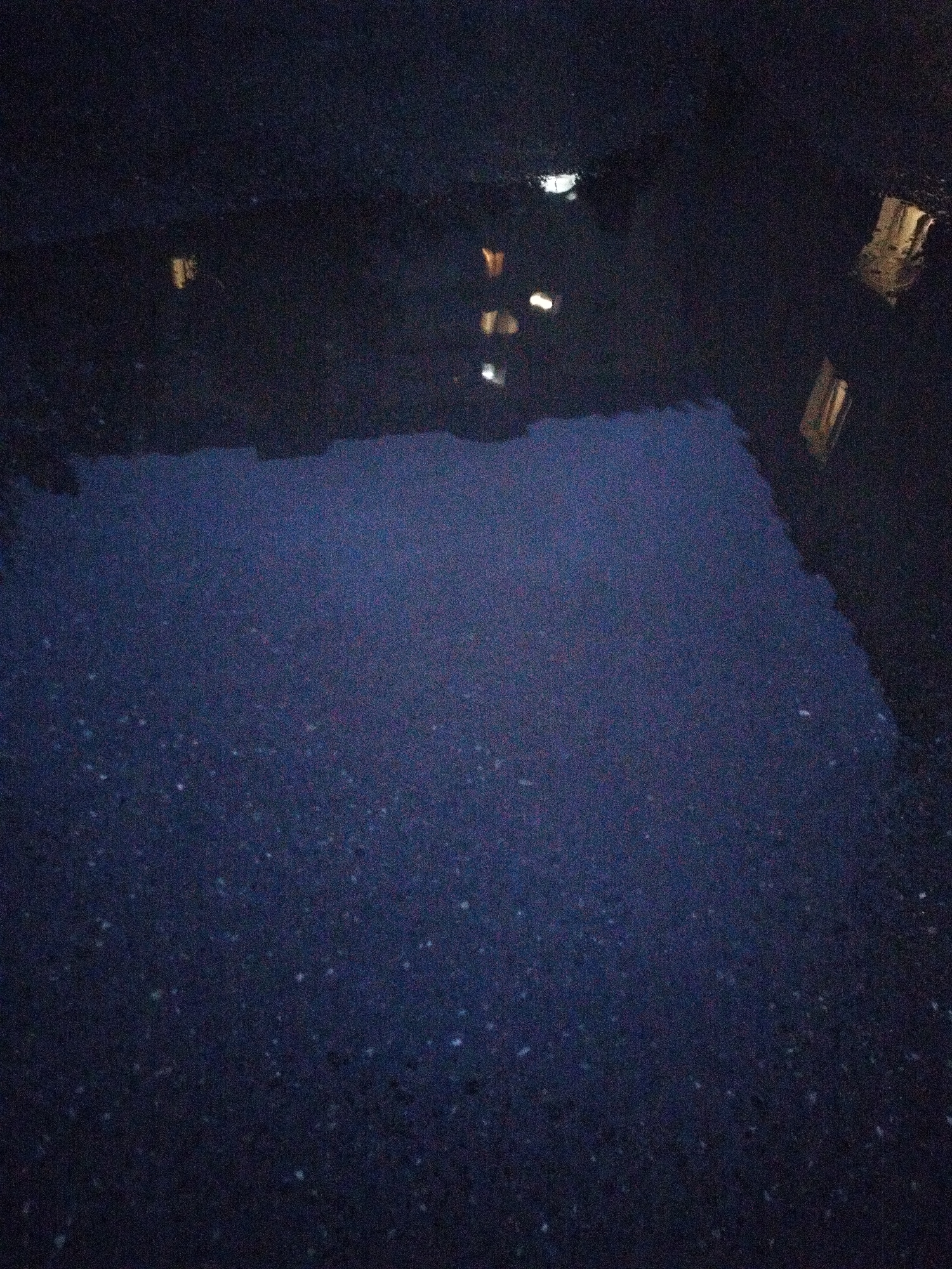 A nighttime photograph shows a puddle of water on asphalt. The water reflects lights from buildings, creating blurry yellow and white shapes. The asphalt itself is dark and textured. The image is grainy and lacks sharp detail, indicating low light conditions. It is not possible to determine the specific location in Surgut, Russia, from this image alone.