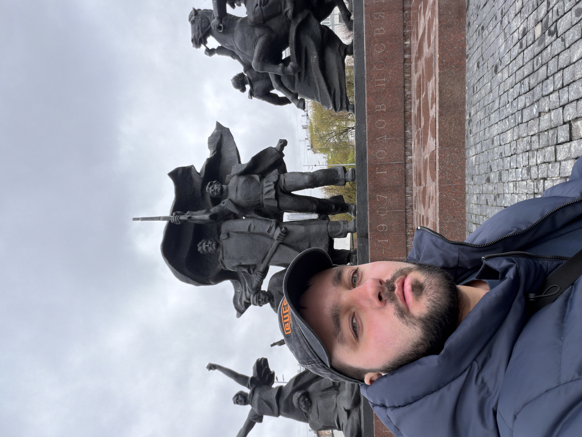 A man takes a selfie, capturing himself and a large, dark-colored monument in Moscow, Russia. The image is oriented vertically, with the man's head and upper torso prominently positioned on the right side. He has a beard and is wearing a dark blue puffer jacket and a dark baseball cap with orange lettering on the front.
Behind him, the monumental sculpture features multiple figures, including individuals on horseback and standing figures, some appearing to hold flags or weapons, all rendered in a dramatic and heroic style. The monument is set upon a substantial reddish-brown stone pedestal. Visible on the side of this pedestal is vertically inscribed Russian text which reads "1907 ГОДОВЩИНА" (1907 ANNIVERSARY). To the right of the monument's base, a section of gray cobblestone pavement is visible. The sky above is uniformly overcast and gray, indicating a cloudy daytime setting. The man looks directly at the camera, posing for the photograph.