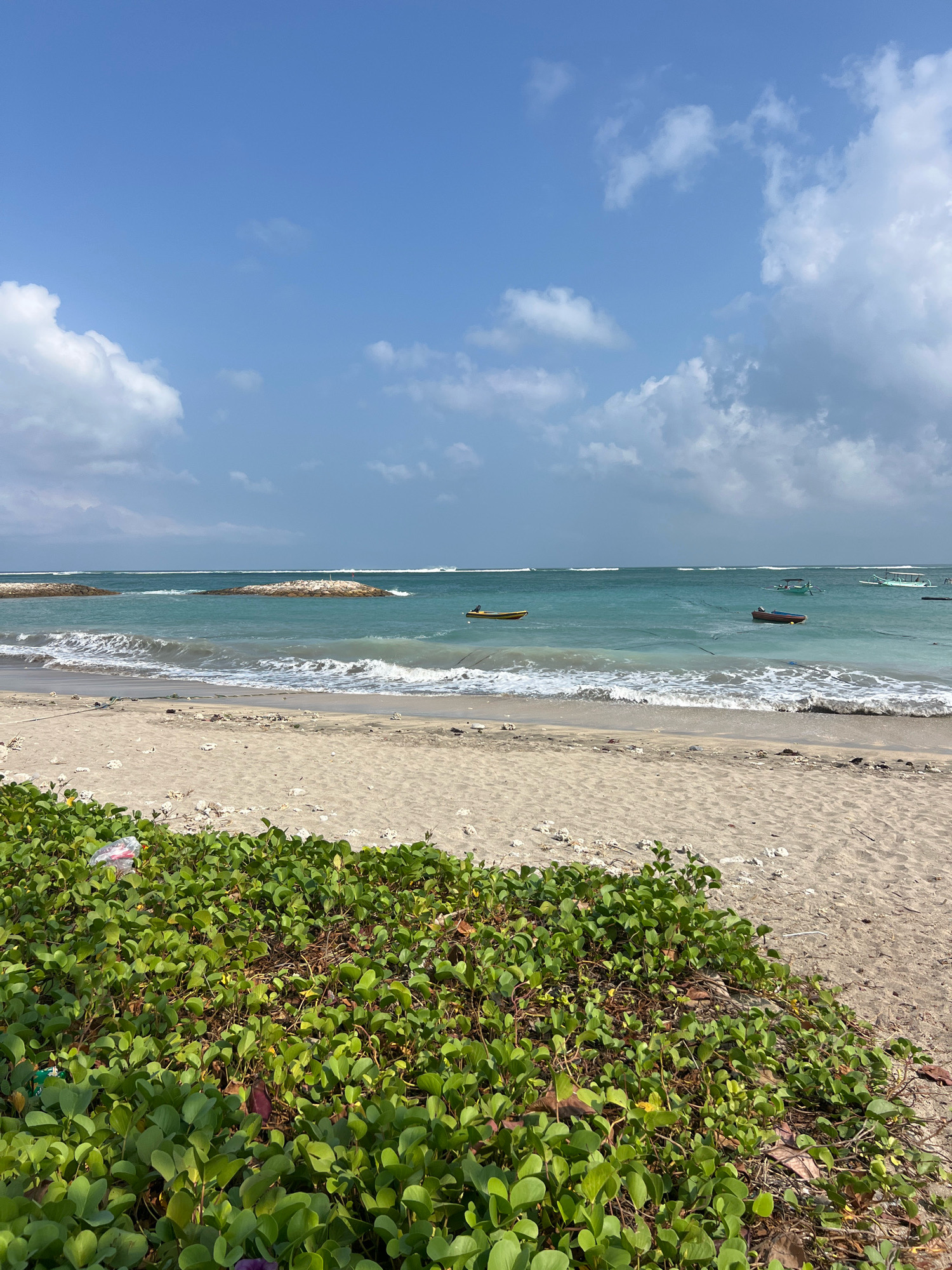 A wide shot of a beach in Kuta, Indonesia. The image captures a sandy shore with lush green vegetation in the foreground. Beyond the beach, the turquoise ocean stretches to the horizon under a partly cloudy blue sky. Several small boats are visible on the water. In the middle ground, two rock formations emerge from the sea, serving as breakwaters. The waves are gently lapping at the shore, and the overall atmosphere is peaceful and serene.