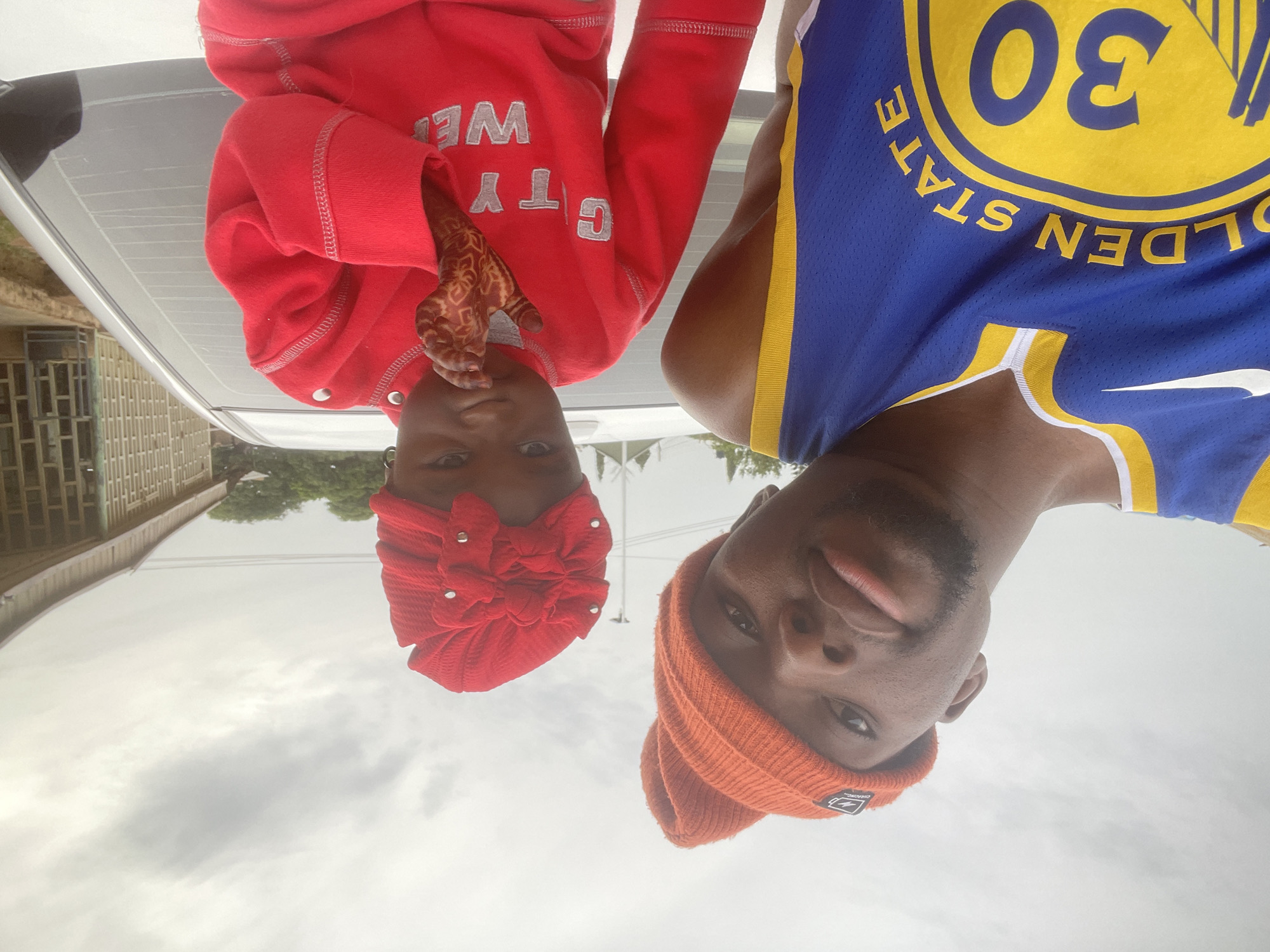 Two people are in the picture. On the left is a child wearing a red head covering and a red sweater with white lettering. The child has henna on their hand. On the right is an adult wearing a blue and yellow Golden State Warriors jersey with the number 30 and an orange beanie.
The background includes a building, a silver car, and a cloudy sky. The image appears to have been taken in Barakin Akawo, Nigeria.