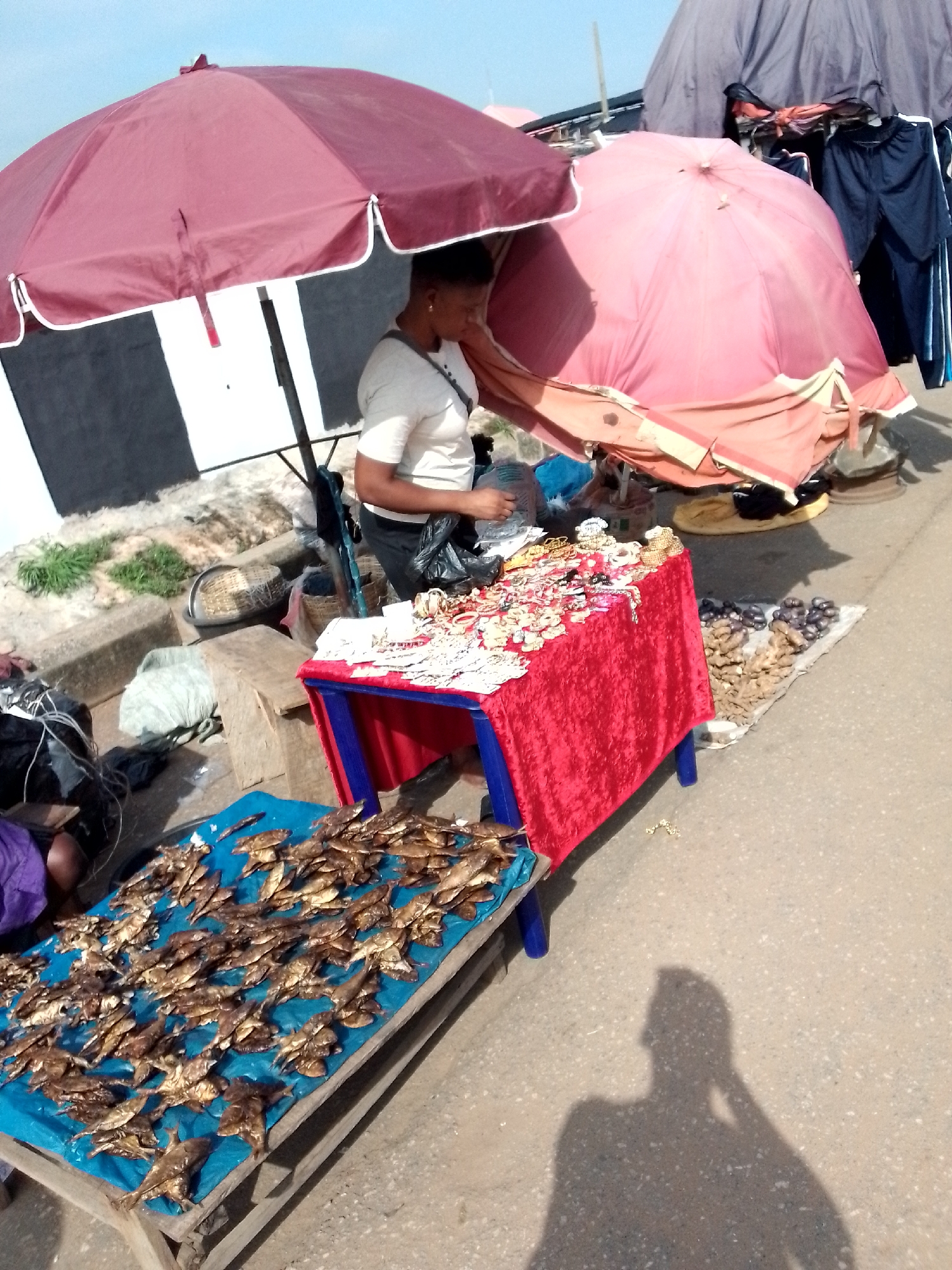 The image captures an outdoor market scene in Nkpor, Nigeria. A woman, wearing a light-colored shirt, is positioned under a large, burgundy umbrella, appearing to be engaged in selling or arranging goods. In front of her, a table draped with a red cloth displays an assortment of jewelry and accessories. To the left of the woman, a table covered with a blue tarp is laden with numerous dried fish, likely for sale. In the background, another large, pink umbrella is visible, along with a structure that appears to be a stall, possibly displaying clothing. The ground is paved and appears to be a street or marketplace. A shadow of a person is cast on the ground in the lower right of the frame, suggesting the photographer or another individual present. The overall atmosphere suggests a typical day at a local Nigerian market.