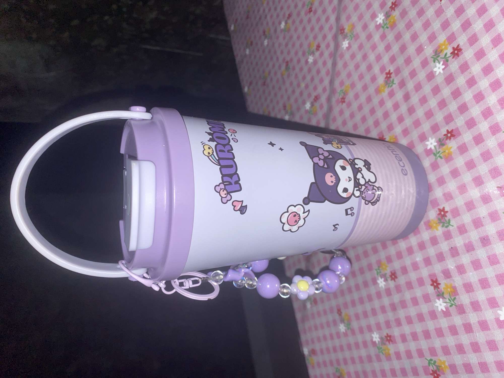 A purple and white tumbler with a white handle is displayed on a pink and white checkered surface with small floral patterns. The tumbler features an illustration of the Sanrio character Kuromi, along with the word "Kuromi" in purple lettering. A beaded charm bracelet, consisting of purple beads and a yellow flower charm, is attached to the tumbler's handle. The background is dark and out of focus. There is no indication of the city of Bontoharu, Indonesia.