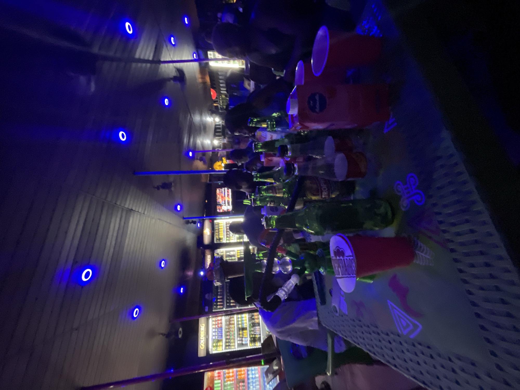 The image depicts an indoor scene at a bar or lounge, with a focus on a table laden with drinks and a hookah pipe. The environment is dimly lit, with prominent blue neon lighting fixtures on the ceiling, creating a vibrant, nocturnal atmosphere. The bar appears to be located in Ikorodu, Nigeria, judging by the branding visible on some of the refrigerators in the background, which display "GOLDBERG" and other beverage logos commonly found in the region.
On the table in the foreground, there are several green glass bottles, some appearing to be beer, along with red disposable cups. A carton of juice or other beverage is also present. A hookah pipe, a centerpiece of the gathering, is positioned prominently. Several individuals are present in the background, their faces partially obscured by the low lighting and the angle of the shot. They appear to be engaged in conversation or socializing. The overall impression is of a lively social gathering at a nightlife establishment.