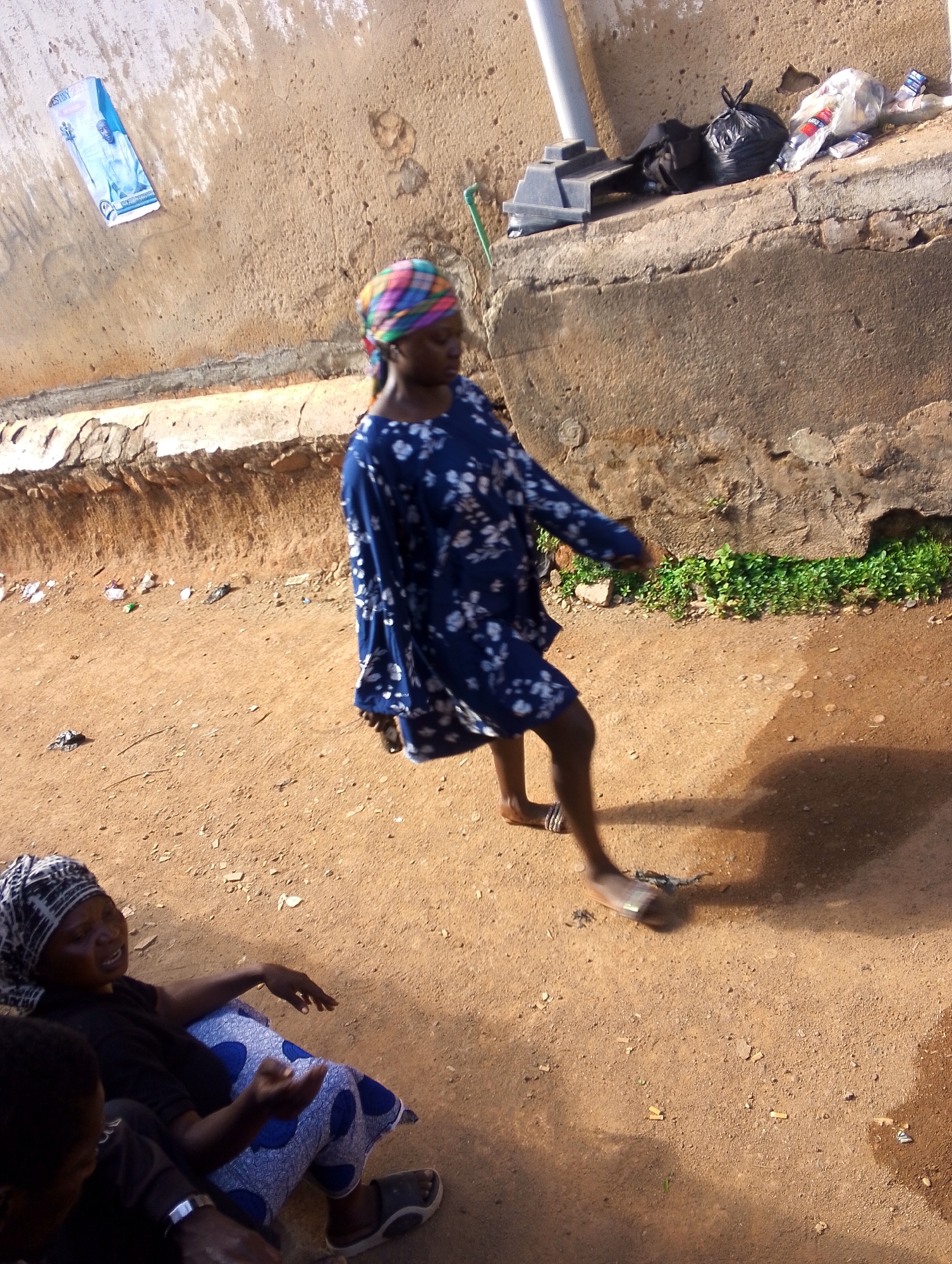 The image, likely taken in the city of Jos, Nigeria, shows a street scene. A young woman in a blue, floral-patterned dress and a colorful headwrap is walking across a dirt area. Her shadow is cast on the ground. To her left and closer to the foreground, a woman is seated on the ground, wearing a black shirt and a blue and white patterned skirt with sandals. Someone else, partially visible and wearing a black shirt and a wristwatch, is sitting near her. In the background, there is a low concrete wall with some trash bags and other debris on top. There is a building with a poster affixed to it. The ground appears sandy and littered with small debris.