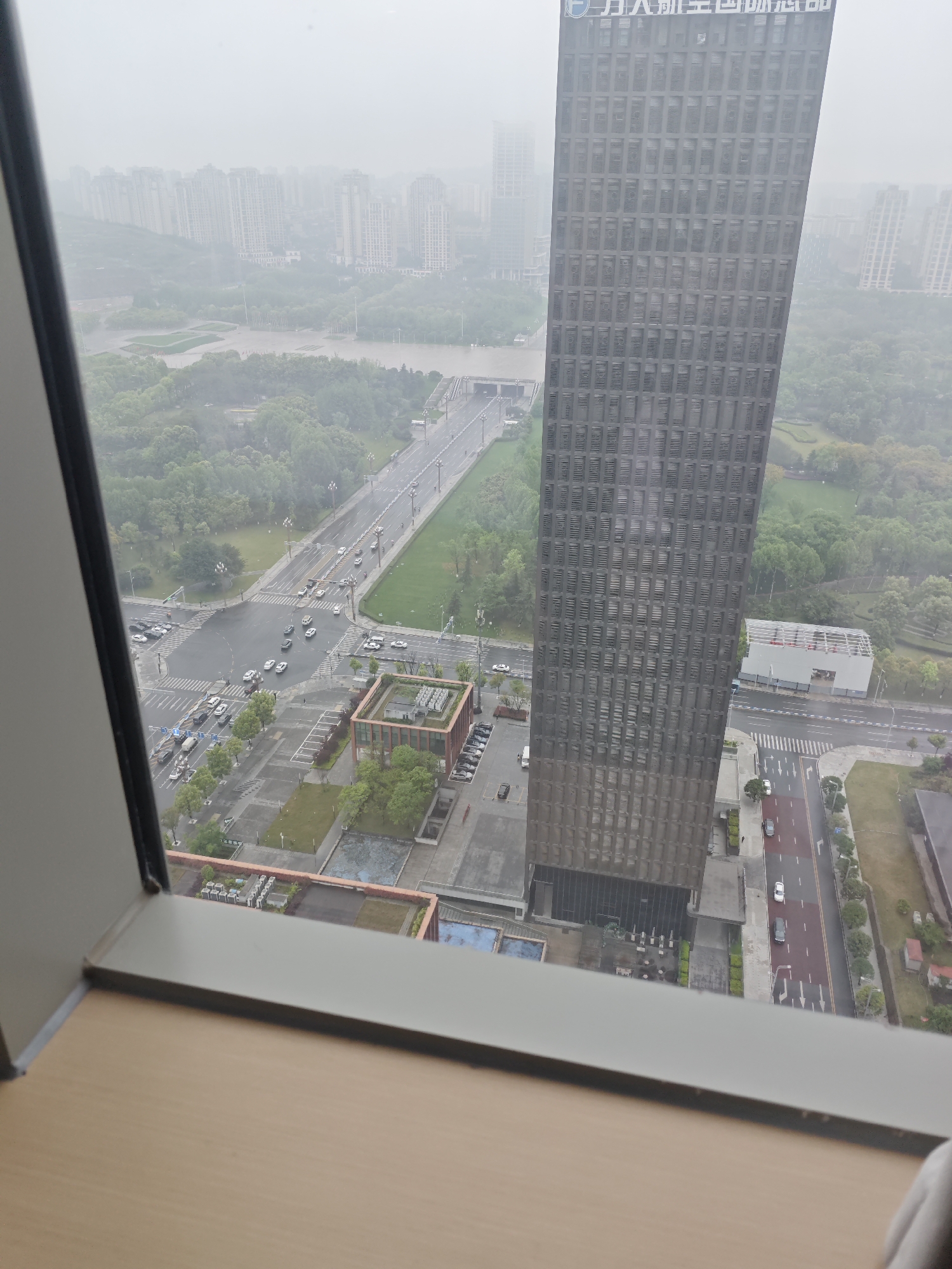 View from a high-rise building in Liangjiang, China, overlooking a cityscape shrouded in heavy mist. In the foreground, a tall, modern skyscraper with a uniform grid of windows dominates the view. Adjacent to it is a multi-story building with a green rooftop. Below, roads with light vehicular traffic intersect. Beyond the roads, a verdant park area stretches towards a river, which is crossed by a bridge. In the far distance, a skyline of numerous high-rise apartment buildings fades into the mist. The photograph is taken through a window with a wooden frame and windowsill.
