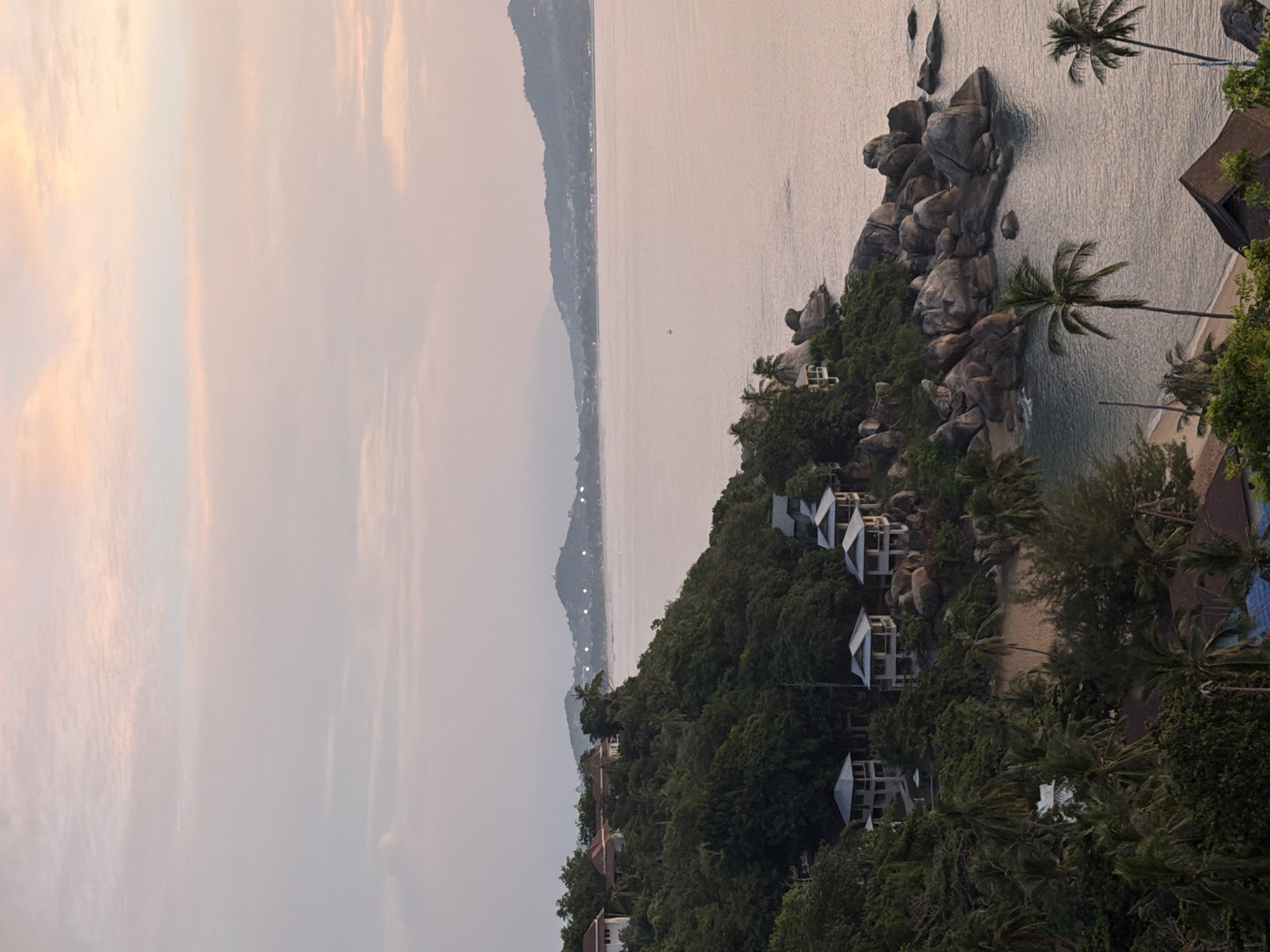 The image depicts a scenic coastal view of Ko Samui District, Thailand, likely during sunset or sunrise. The foreground features lush green trees and palm trees framing several modern villas with white roofs, nestled on a hilly terrain overlooking the ocean. The villas appear to be residential or resort properties.
The middle ground is dominated by the calm, expansive sea, reflecting the soft hues of the sky. Rocky outcrops with some vegetation emerge from the water along the coastline. In the distance, the opposite shore of Ko Samui is visible, characterized by rolling hills and mountains, with scattered lights indicating human settlements or development. The sky is painted with delicate shades of pink, orange, and pale blue, indicative of a twilight atmosphere. There are no people visible in the frame. The overall impression is one of serene tropical beauty and peaceful seclusion.