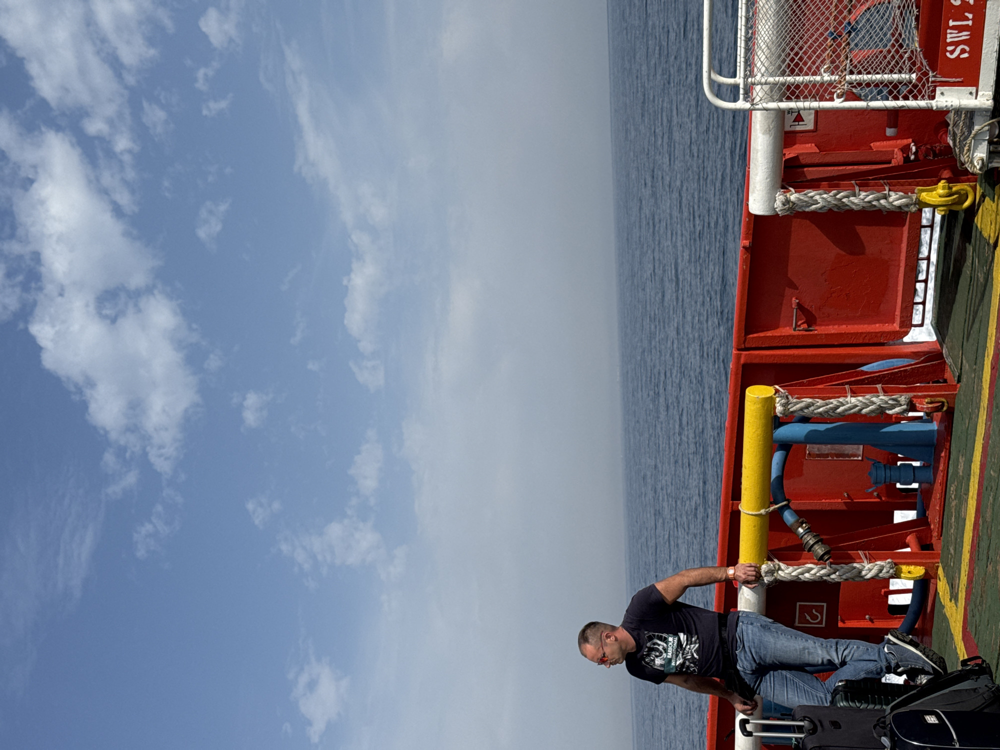 A man wearing a black t-shirt and blue jeans stands on the deck of a ferry, leaning against the railing. He is wearing sunglasses and a watch. Two suitcases are beside him. The ferry deck is red with yellow and green markings. Thick ropes are tied to the railing. The sea is visible in the background, with a hazy horizon. The sky is blue with scattered white clouds. The image appears to have been taken in Marina di Ravenna, Italy, as indicated by the context.
