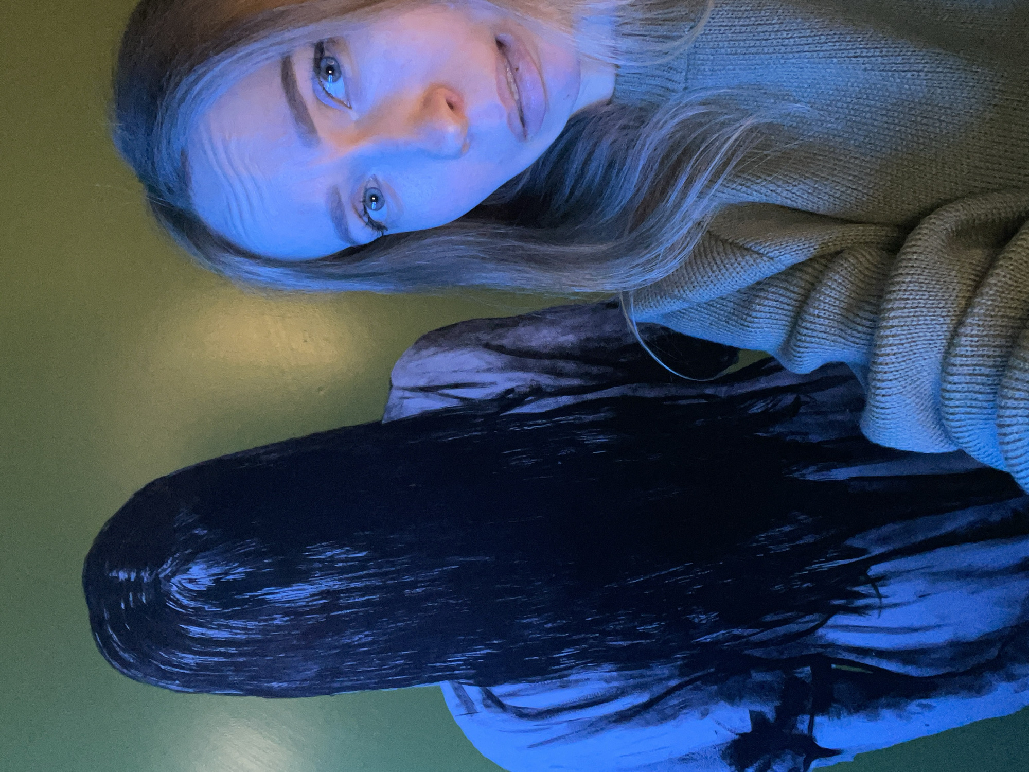 A photograph shows a young woman with blonde hair wearing a light green sweater. Next to her is what appears to be a glossy, black wig laying on a green surface. The photograph has a blue color cast. The location is not discernible and therefore cannot be placed within Perm, Russia or any other specific location.