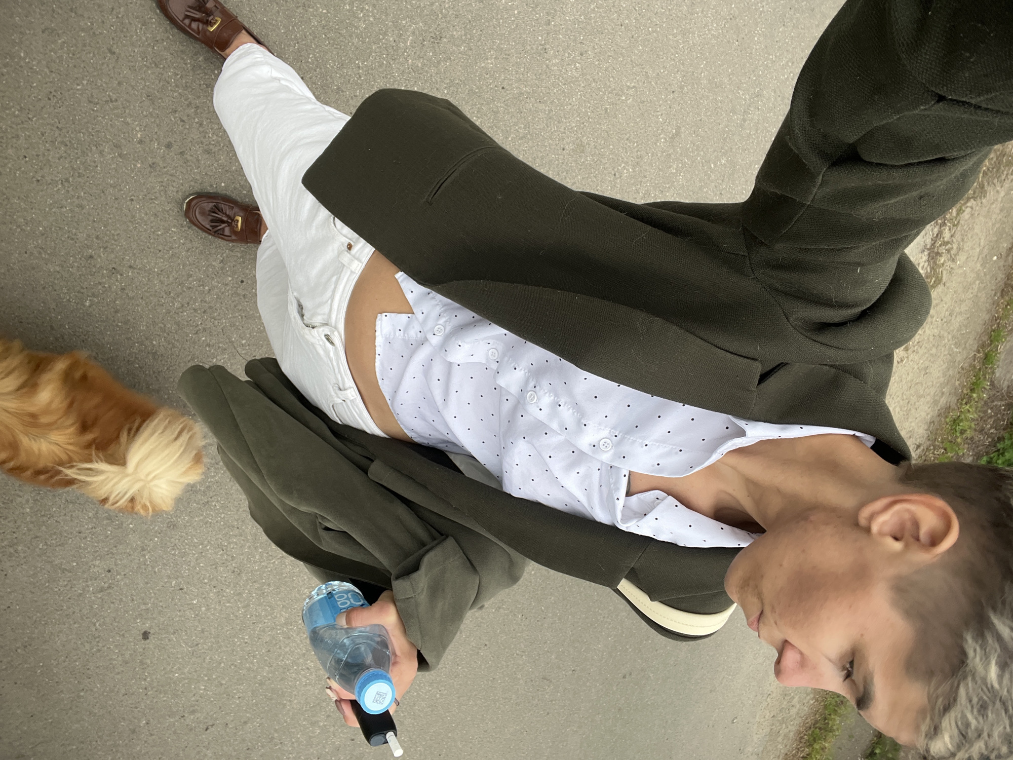 A medium, angled shot depicts a person on a paved surface, possibly a sidewalk or path, holding a plastic water bottle in their left hand and what appears to be an electronic cigarette in the same hand. A tan-colored dog is partially visible at the left edge of the frame.
The person is wearing white jeans, brown loafers, a white blouse with black polka dots, and an olive-green blazer. The person's face, oriented to the right of the frame, displays short, light-colored hair.
The setting appears to be outdoors, with green foliage visible at the right edge of the frame. The location is uncertain, and there is no information indicating the city of Boguchar, Russia.