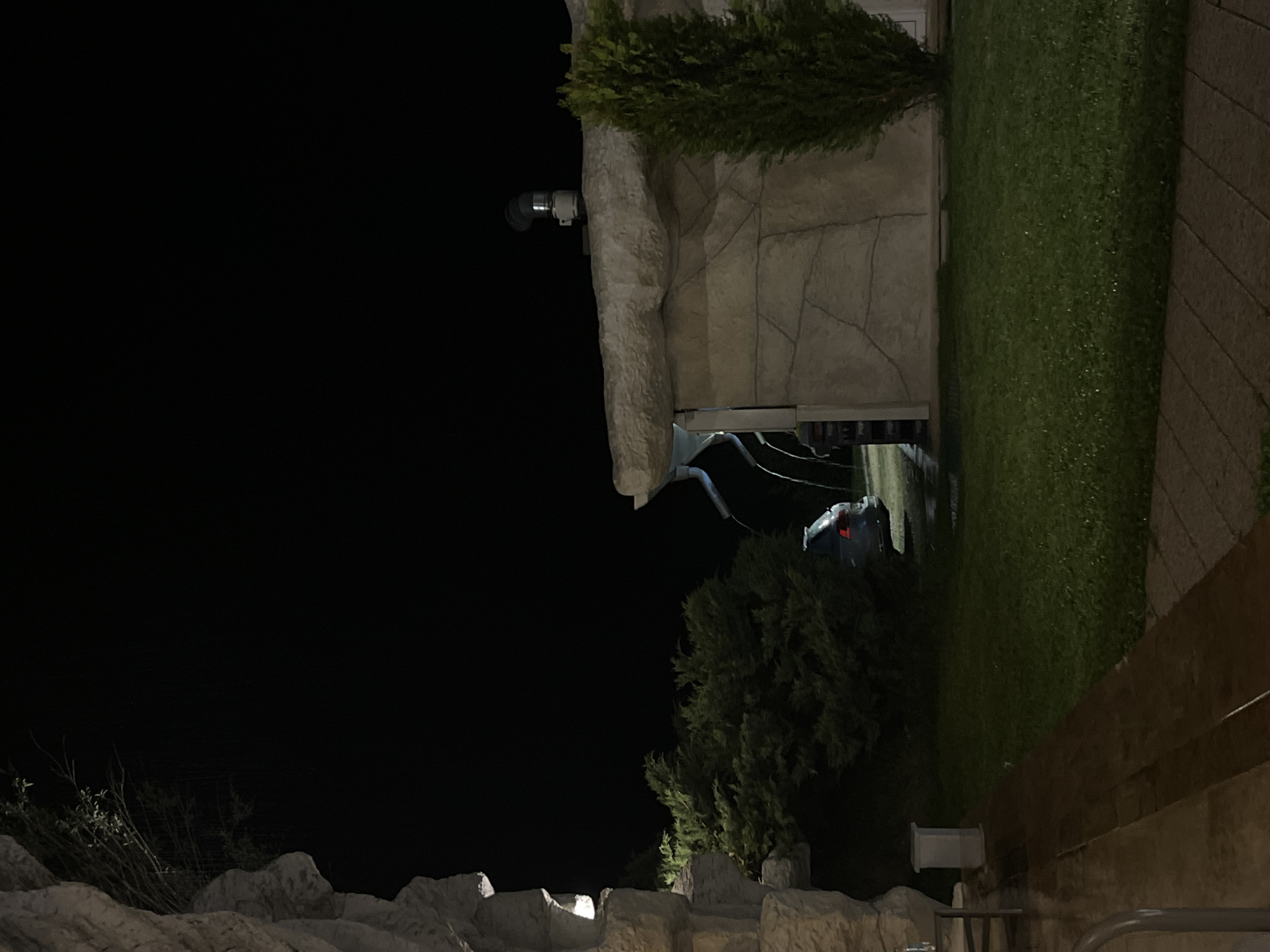 A night scene in Koprivlen, Bulgaria. The image shows a building with a stone facade, overgrown with foliage, and illuminated by an unseen light source. A security camera is mounted on the building. A dark, seemingly empty street is visible through an opening in the building. The lower portion of the image shows a stone structure with vegetation growing around it. The rest of the image is mostly dark.