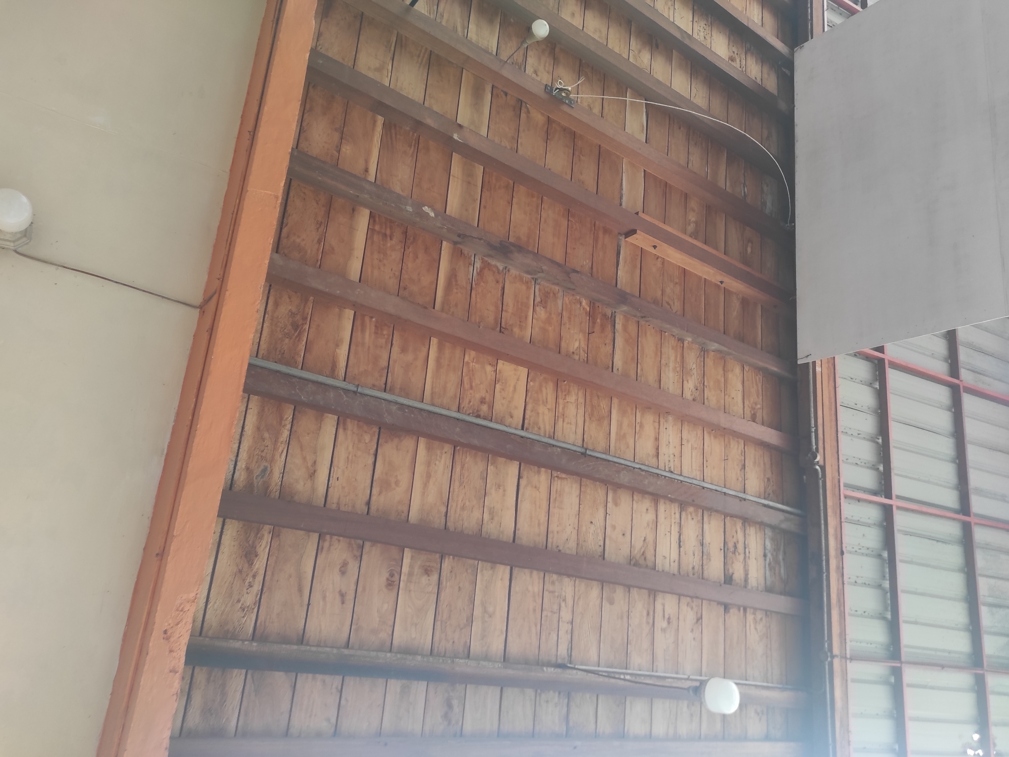 The image displays an upward view of an architectural structure, likely a ceiling or high wall, set in Manetunong, Indonesia. The dominant feature is a rustic wooden surface composed of numerous vertical light-brown wooden planks. These planks are horizontally traversed and supported by several darker, unpainted or stained wooden beams.
Running along some of these wooden beams are grey metal conduits or pipes, suggesting electrical or utility lines. Two spherical white light fixtures are visible, one in the upper-middle section and another towards the lower right, both emitting a soft glow and attached to the wooden structure. A thin white wire arches across the upper right section of the wooden ceiling.
To the left, a thick, vertical beam painted in a vibrant orange hue acts as a structural boundary, separating the wooden ceiling from a plain, light-colored wall surface. On the far right, a large, rectangular white panel hangs vertically, partially obscuring a background structure. Beyond this panel, a framework of reddish-brown metal beams supports what appears to be a section of corrugated metal siding or another wall element.
The scene is brightly lit, indicative of daytime. There are no people, explicit activities, or visible text within the frame. The overall impression is one of utilitarian construction, emphasizing natural wood and functional elements.