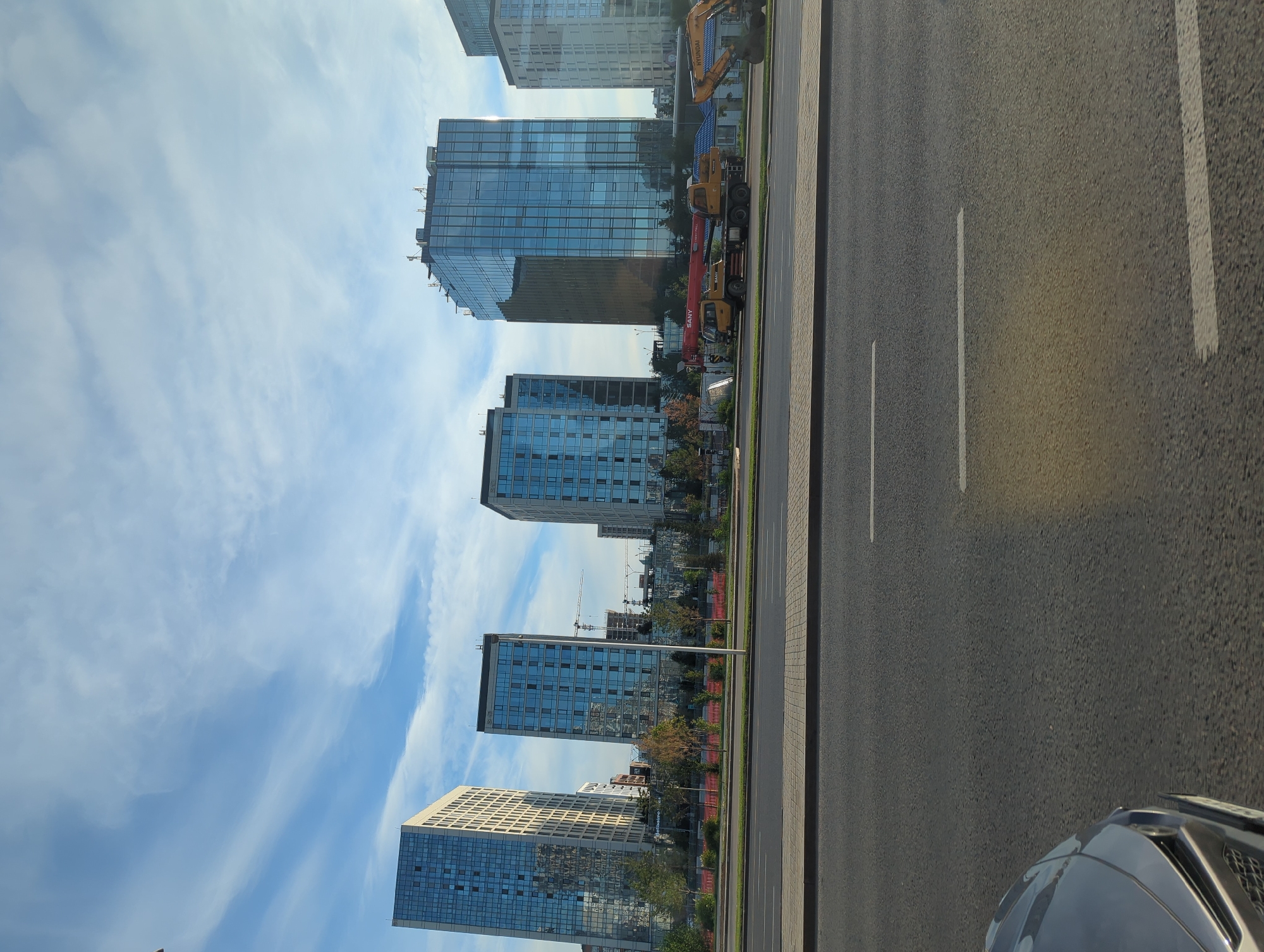 The image depicts a cityscape in Astana, Kazakhstan, featuring a row of modern glass-facade buildings under a partially cloudy sky. In the foreground, a paved road with white lane markings occupies the lower half of the frame, with the hood of a silver car visible on the left. Behind the road is a green strip with trees and shrubs, separating it from the buildings. Construction activity is evident behind the buildings, with cranes and partially constructed structures visible. A yellow crane truck with "SANY" branding and a yellow excavator with "HYUNDAI" branding are parked near the buildings. The overall scene suggests a developed urban area with ongoing construction and typical city infrastructure.