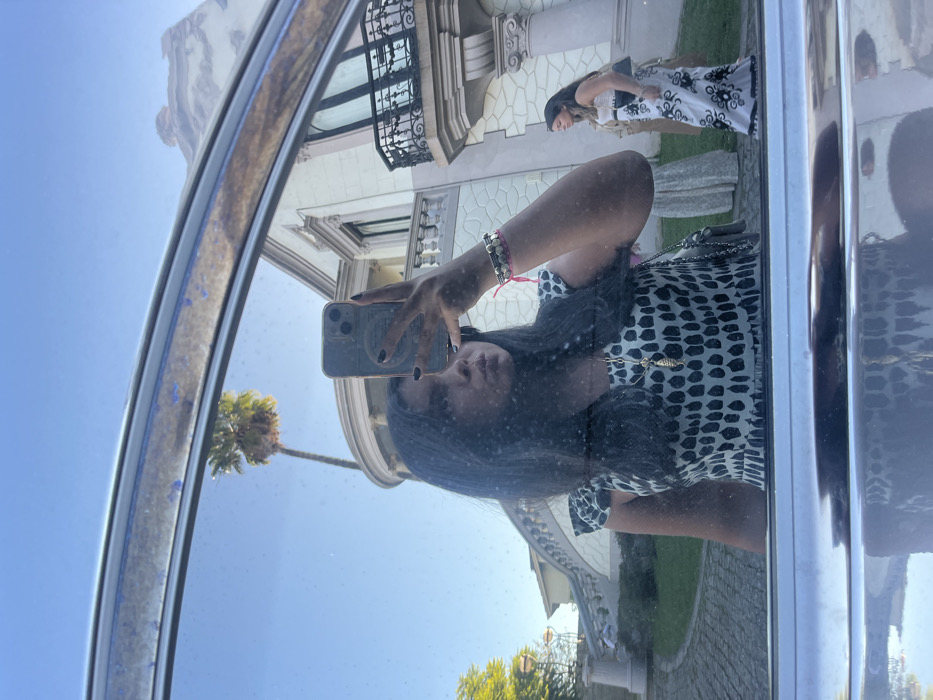 The image captures a mirror selfie taken from within a vehicle, reflected on a slightly smudged window or mirror surface. In the foreground, a woman with long dark hair, dark nail polish, and a black and white patterned top or dress, holds a light-colored smartphone with a circular grip in her right hand to capture the reflection. She wears multiple bracelets, including a pink string and beaded styles, on her left wrist, and a gold-colored necklace with an elongated pendant. Her lips are pursed.
The background, visible through the reflection, features a grand, ornate building situated in Lordelo do Ouro e Massarelos, Portugal. This multi-story structure is light-colored, displaying Neoclassical or Romantic architectural characteristics, including prominent columns, decorative cornices, and a sweeping, curved exterior stone staircase with dark railings on its left side leading to an upper-level entrance. A dark wrought-iron balcony adorns an upper window on the right side of the building. To the left, a tall palm tree stands against a clear blue sky, with additional green foliage visible further left. The ground in front of the building is paved with cobblestones. Two other women are visible in the background: one on the right wears a long white dress with a distinct black pattern and carries a dark clutch bag; the other, partially obscured behind the first and near the staircase, wears a light-colored long dress. The scene is illuminated by bright daylight.