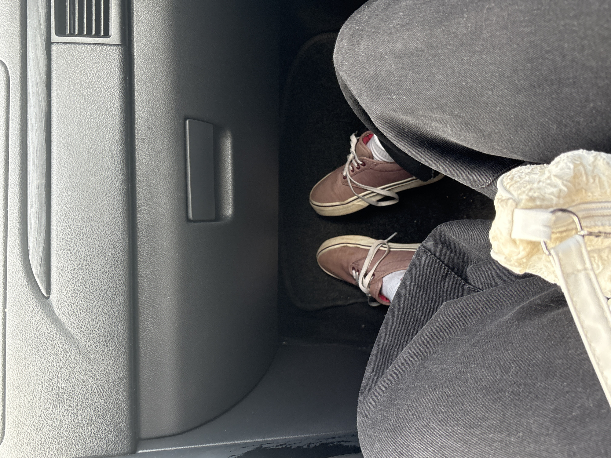 The image shows the interior of a vehicle, possibly a car, with a focus on the passenger side. The passenger is wearing black pants and brown sneakers with white laces. A small white purse with a zipper is visible near the passenger's leg. Part of the dashboard, with an air vent and a recessed handle, is on the left side of the frame. The location, Chaikovsky, Russia, cannot be confirmed from the visual information provided.