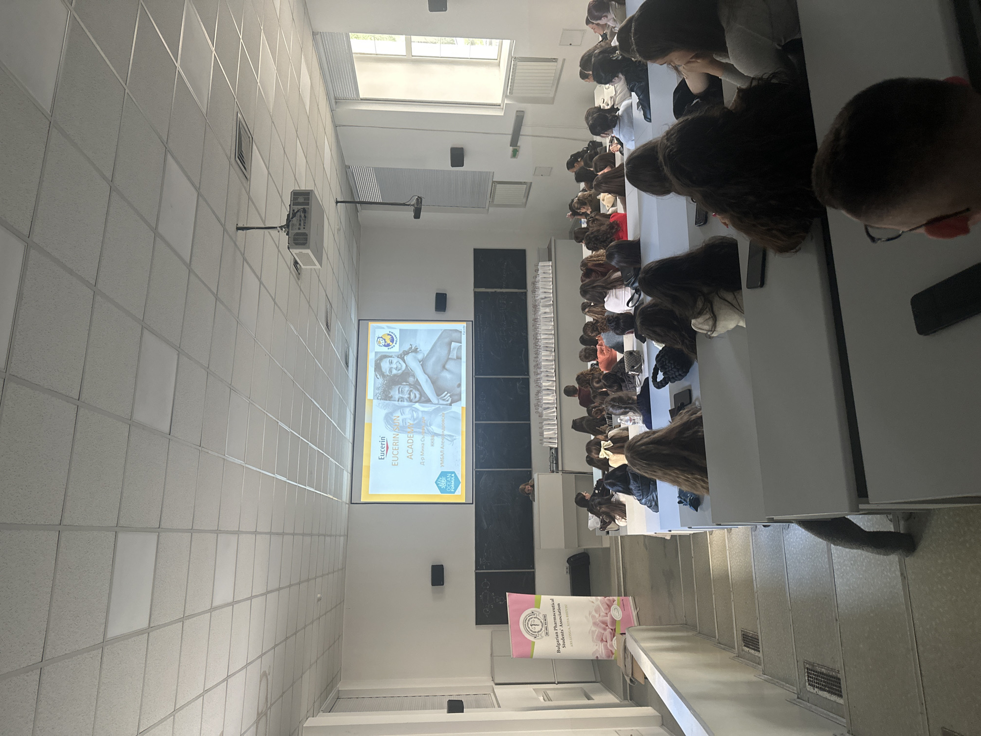 The image shows an indoor scene of what appears to be a lecture hall or classroom in Sofia, Bulgaria. A group of students is seated at rows of white tables, facing a projection screen where an advertisement for the skincare brand Eucerin is displayed. The banner for the "Bulgarian Pharmaceutical Students' Association" is standing to the right of the screen. The environment suggests an educational setting, likely within a university or professional development context.