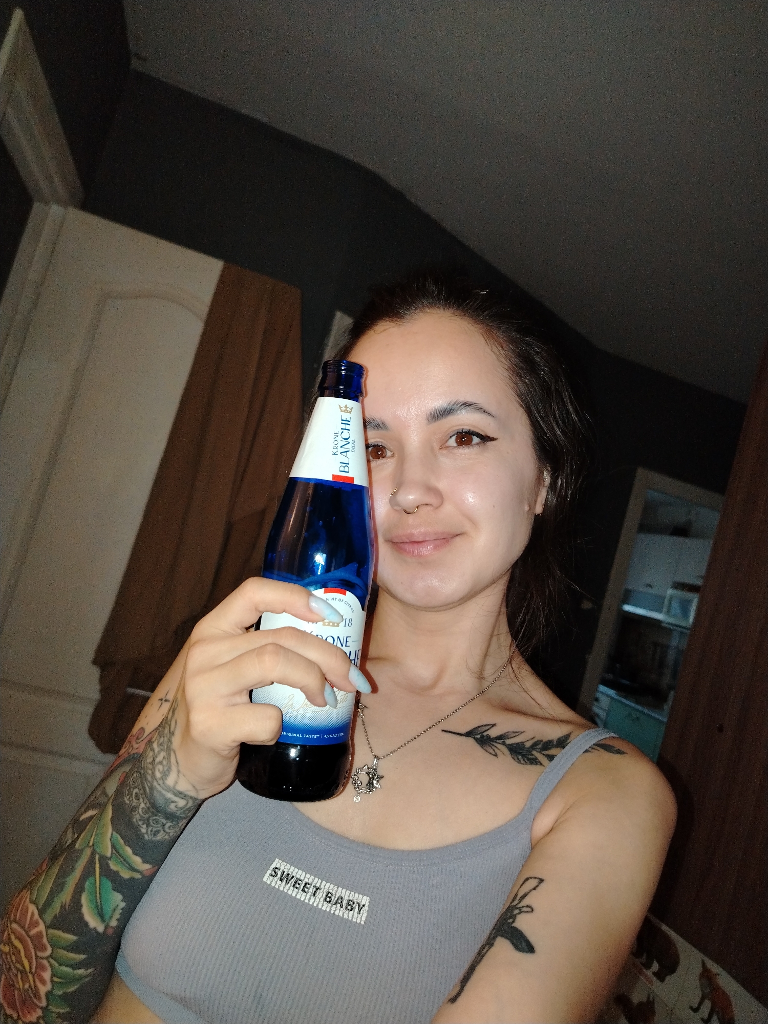 The image features a young woman posing for a selfie indoors in Noviy, Russia. She appears to be in her 20s or 30s, with dark hair pulled back, brown eyes, winged eyeliner, and a septum piercing. Her expression is a gentle, slight smile.
She is wearing a light grey ribbed tank top that has a white rectangular patch on the chest with the text "SWEET BABY" printed in black capital letters. Her left arm is heavily tattooed with a large sleeve design featuring vibrant floral patterns and dark shading. She also has a tattoo of a leafy branch on her left shoulder/collarbone area and another smaller, indistinct tattoo on her right upper arm.
The woman is holding a blue glass bottle with a white label in her right hand. Her fingernails are painted a light blue. Visible text on the bottle's label includes "KRONE BLANCHE BIERE" along with a crown logo, "MINT OF CITRUS," "ORIGINAL TASTE," and "4.5% ALC/VOL."
The background is dimly lit, suggesting an indoor setting, possibly in the evening or night. Dark walls are visible, along with parts of a white door or doorframe on the left. On the right, a brown piece of furniture or fabric is present, and a subtle reflection in what appears to be a mirror shows indistinct shapes of a room beyond.