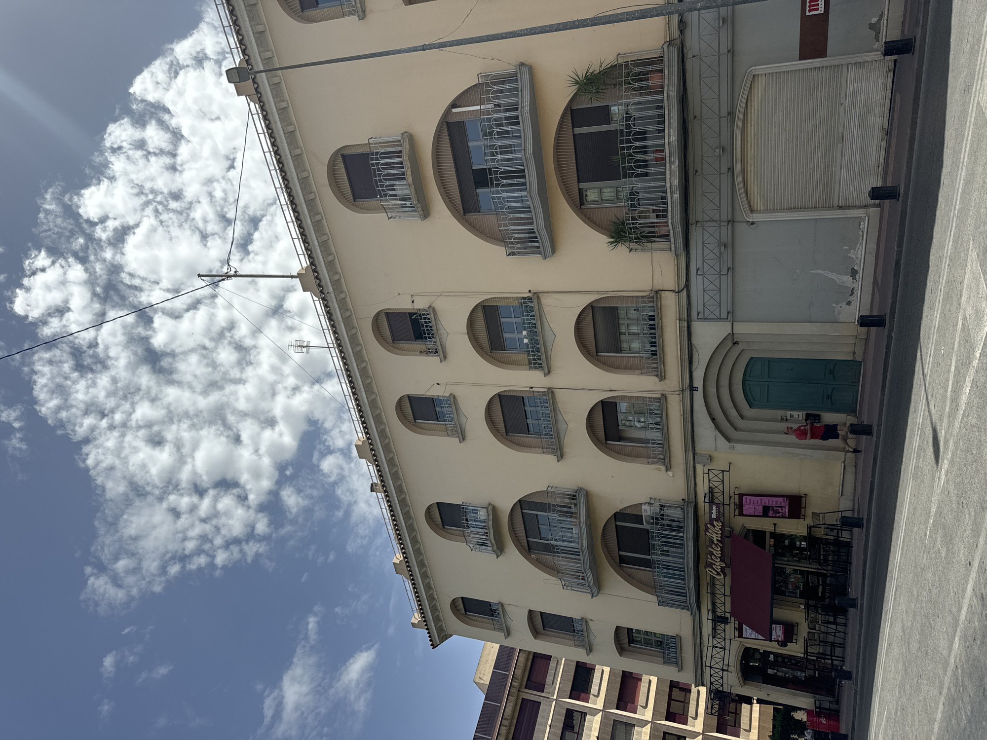 This is a photograph of a building with a light yellow facade and numerous arched windows, each with a small balcony. The windows on the upper floors are arranged in two rows, with four windows on the top row and five on the second. The building has a cornice running along the top. The sky above is blue with fluffy white clouds, suggesting it is daytime and the weather is pleasant.
The bottom of the building has a storefront with tables and chairs set up outside, suggesting it is a cafe or restaurant. A red awning is extended over the seating area. The name of the establishment, "Café de Alba," is visible on the facade. A closed security shutter is visible on the right side of the building. A person in a red shirt and blue shorts is standing near a turquoise door on the right side of the building. Another person is partially visible in red near a trash can.
The photograph is taken at an angle, with the building and sky occupying the majority of the frame. The lighting indicates a sunny day. There are electrical wires running diagonally across the upper left portion of the image. The architectural style of the building, with its arched windows and balconies, is characteristic of European cities. The overall scene conveys a sense of a typical urban street in a warm climate.