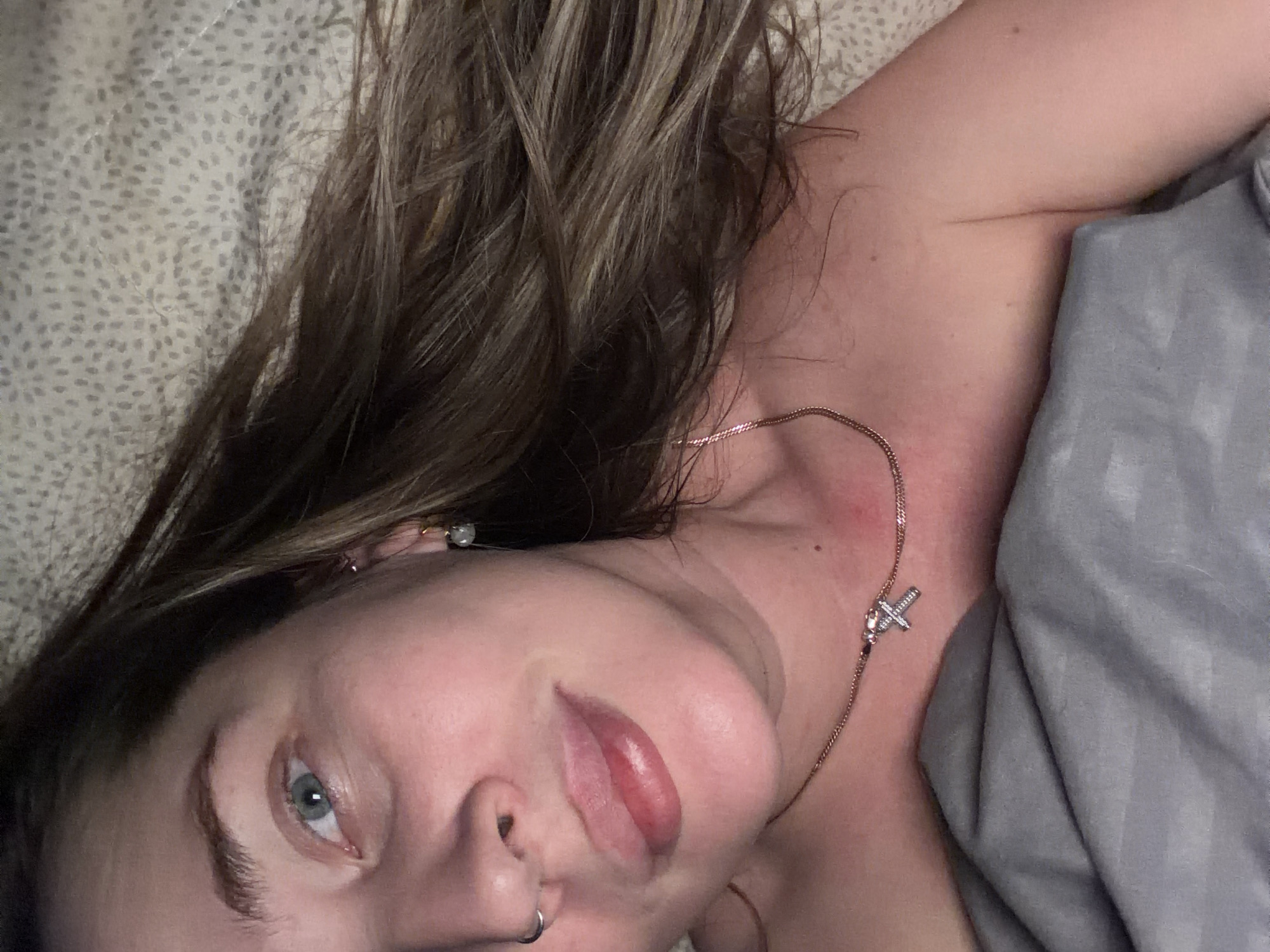 A close-up, overhead view depicts a female subject lying supine in an indoor setting. Her head is slightly turned, and she looks directly towards the camera. The visible features include her left eye, which is light in color, a nose with a silver-toned septum ring, and full, slightly parted lips exhibiting a faint smile. Her long, wavy, brown hair with lighter highlights is spread out behind her head, resting on the background surface. She wears a gold-toned chain necklace with a small, cross-shaped pendant adorned with tiny reflective elements, and a small hoop earring with a light-colored dangling stone in her visible left ear. A grey fabric, consistent with a blanket or duvet, covers her lower visible torso, while her upper chest and right shoulder are exposed. A faint reddish mark is present on the skin below her neck. The background consists of a light-colored fabric patterned with small, dark, irregular shapes, suggesting bed linens. The lighting is soft and ambient. No specific visual cues are present in the image to determine the precise location within the city of Chaikovsky, Russia, beyond an indoor, private environment.