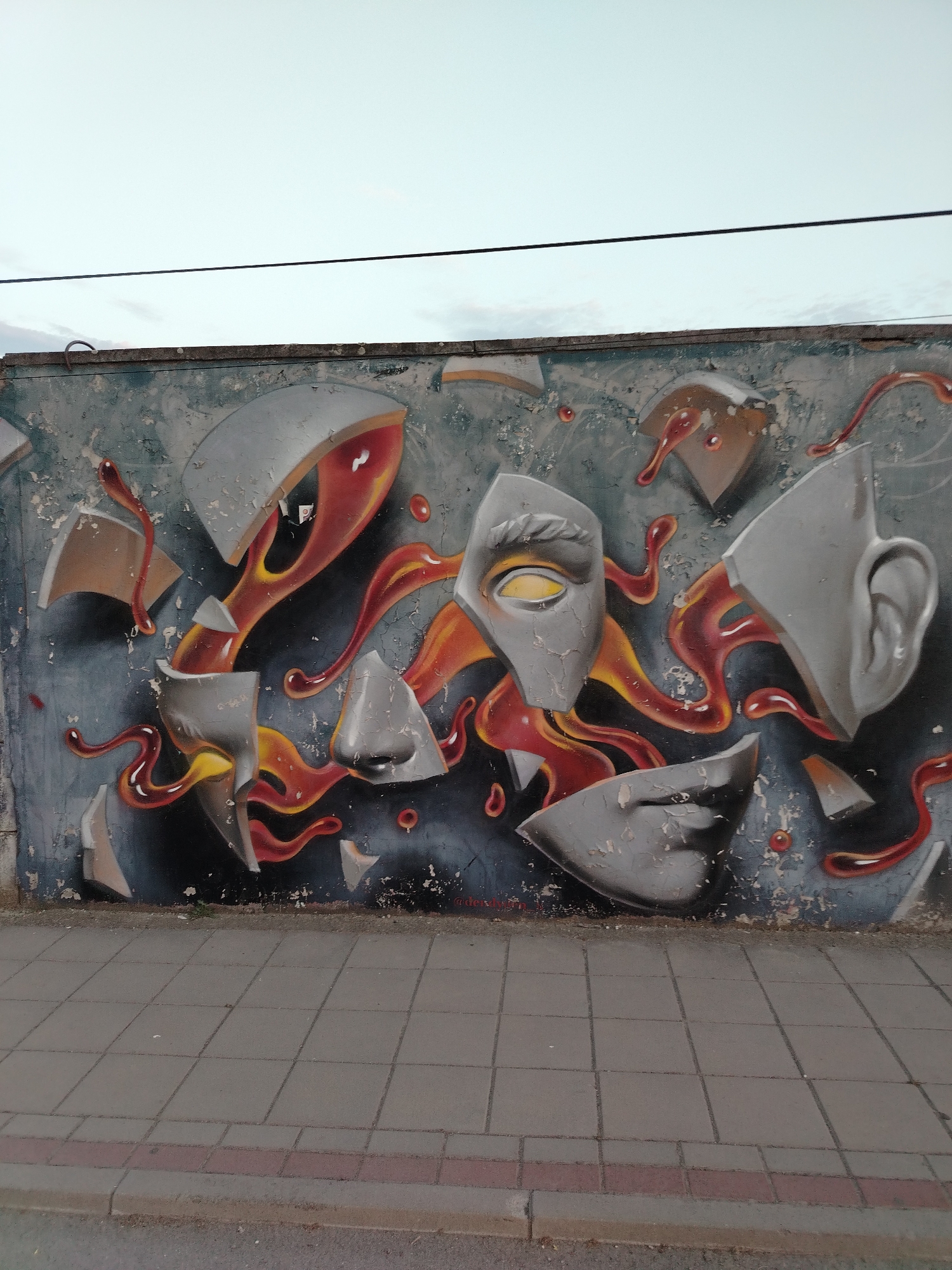 The image shows a section of a graffiti-covered wall, likely located in Novi Sad, Serbia. The graffiti depicts a surreal, fragmented face or mask, with various pieces seemingly breaking apart and spilling a viscous, reddish-orange liquid. The fragments include an eye with a yellow iris, a nose, and parts of a mouth and ear. The background of the graffiti is a dark, textured gray, with lighter gray and white accents, and the "liquid" has highlights of yellow and orange, suggesting a glossy or molten texture. The wall itself appears weathered and shows signs of peeling paint and cracks. In front of the wall, there is a paved sidewalk made of grey square tiles, with a red-bordered section closer to the street. A grey, unpaved strip of ground is visible between the sidewalk and the bottom edge of the image. Above the wall, a pale, overcast sky is visible, with a dark electrical wire stretching horizontally across the upper portion of the frame. No people are present in the image.