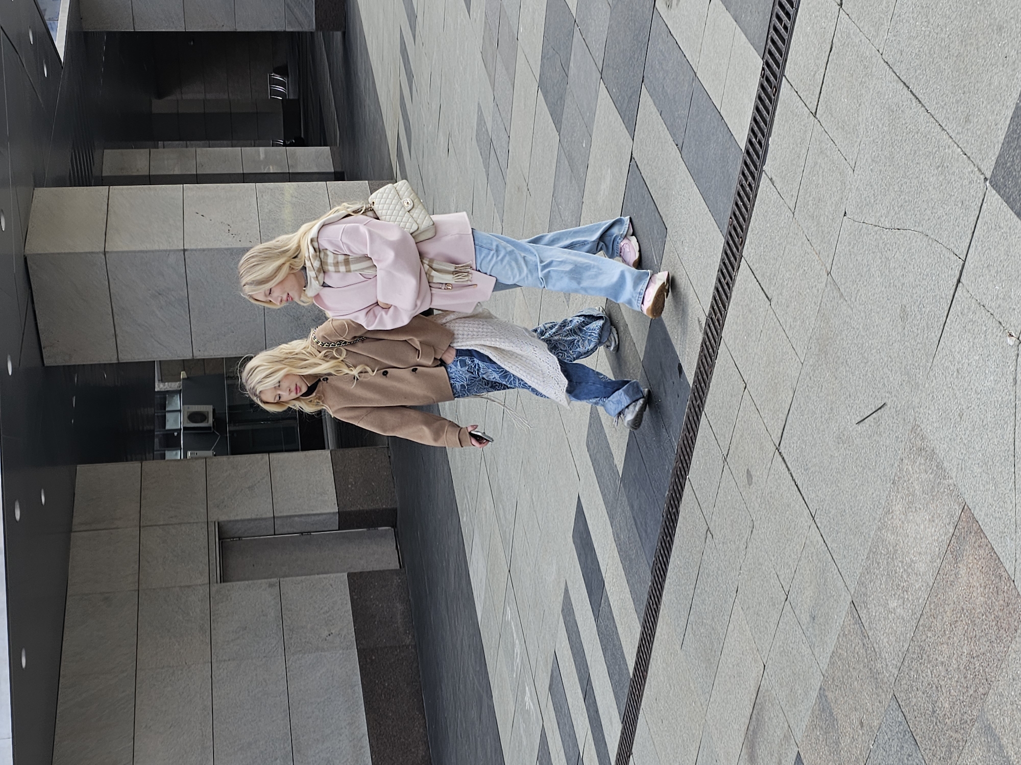 Two blonde women walk side-by-side on a paved surface near a building in Moscow, Russia. The woman closer to the camera wears a tan coat, blue jeans, and carries a cell phone. The other woman wears a pink jacket, blue jeans, and a white purse. The surrounding environment includes modern architecture and patterned pavement.