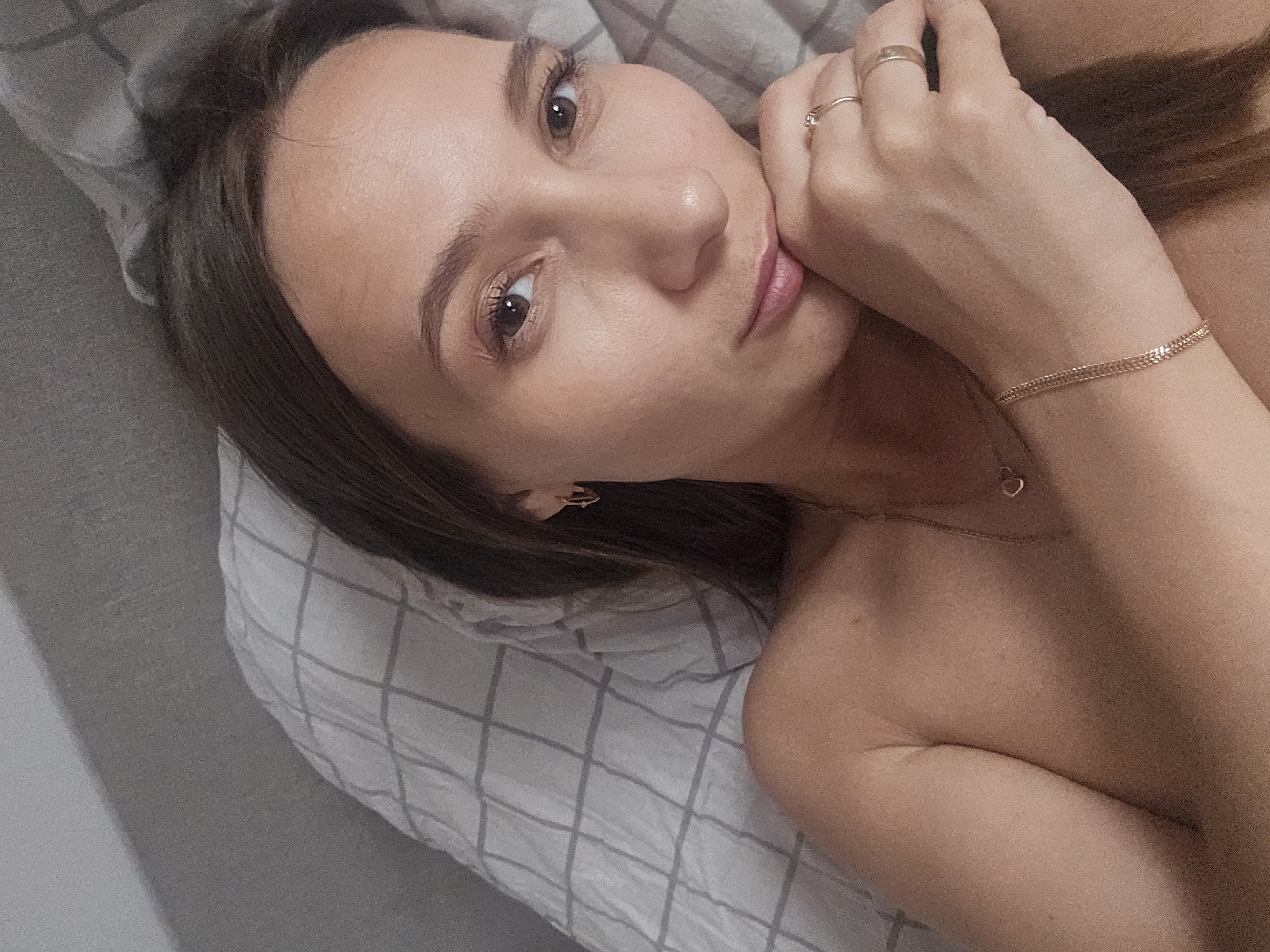 A young woman is lying down, resting her face on her hand. She has brown hair, light skin, and is wearing a gold necklace, bracelet, and rings. The background includes a gray pillow with a white grid pattern and a gray wall. There is no information available about the location in Chaikovsky, Russia.