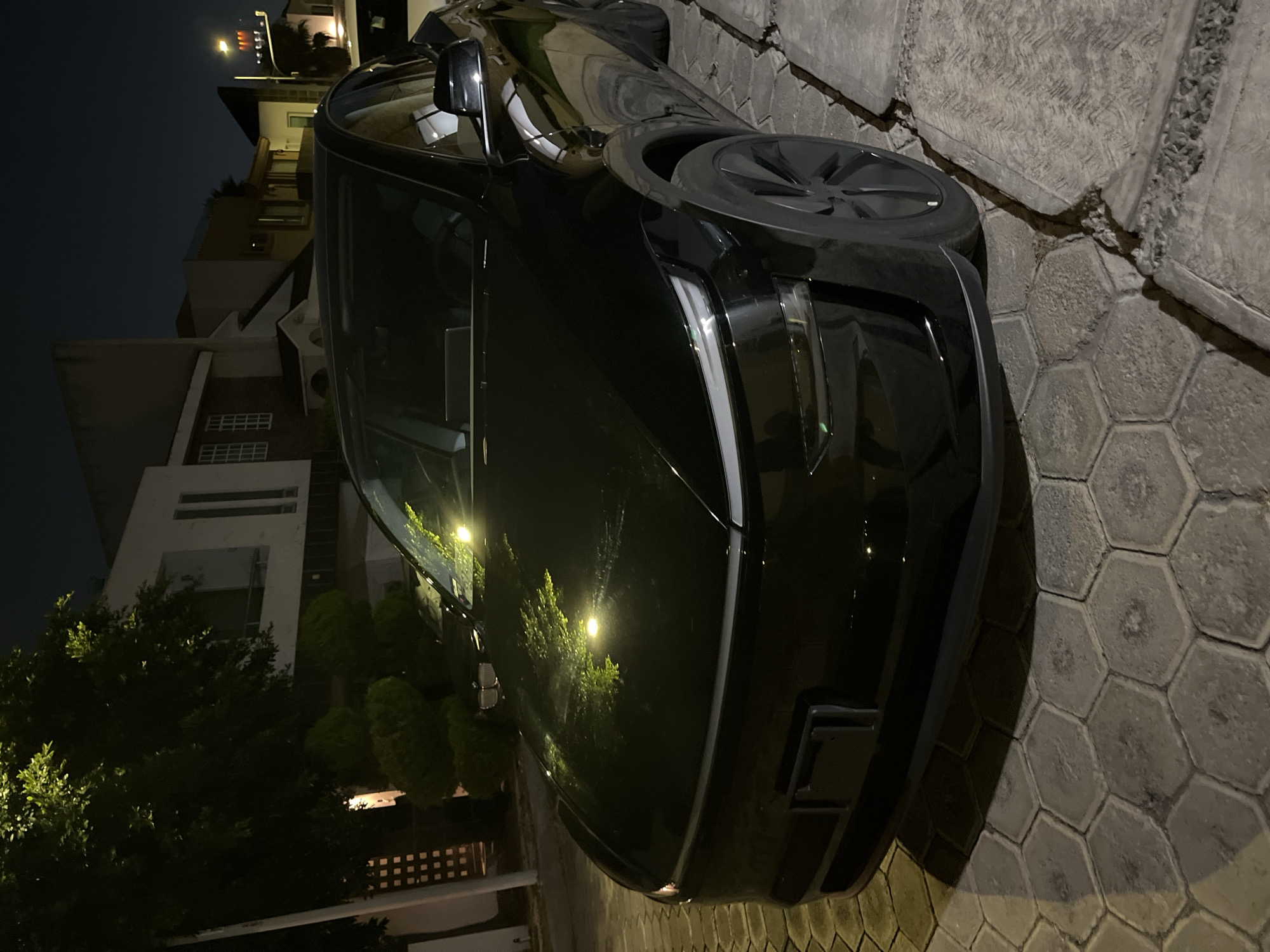 A black car is parked on a street in San Bernardino Tlaxcalancingo, Mexico. The car is parked on a paved street of hexagon shaped gray pavers, with a raised section of concrete running along the edge. The car is reflecting the surrounding foliage. In the background are buildings, trees, and street lights. The sky is dark.