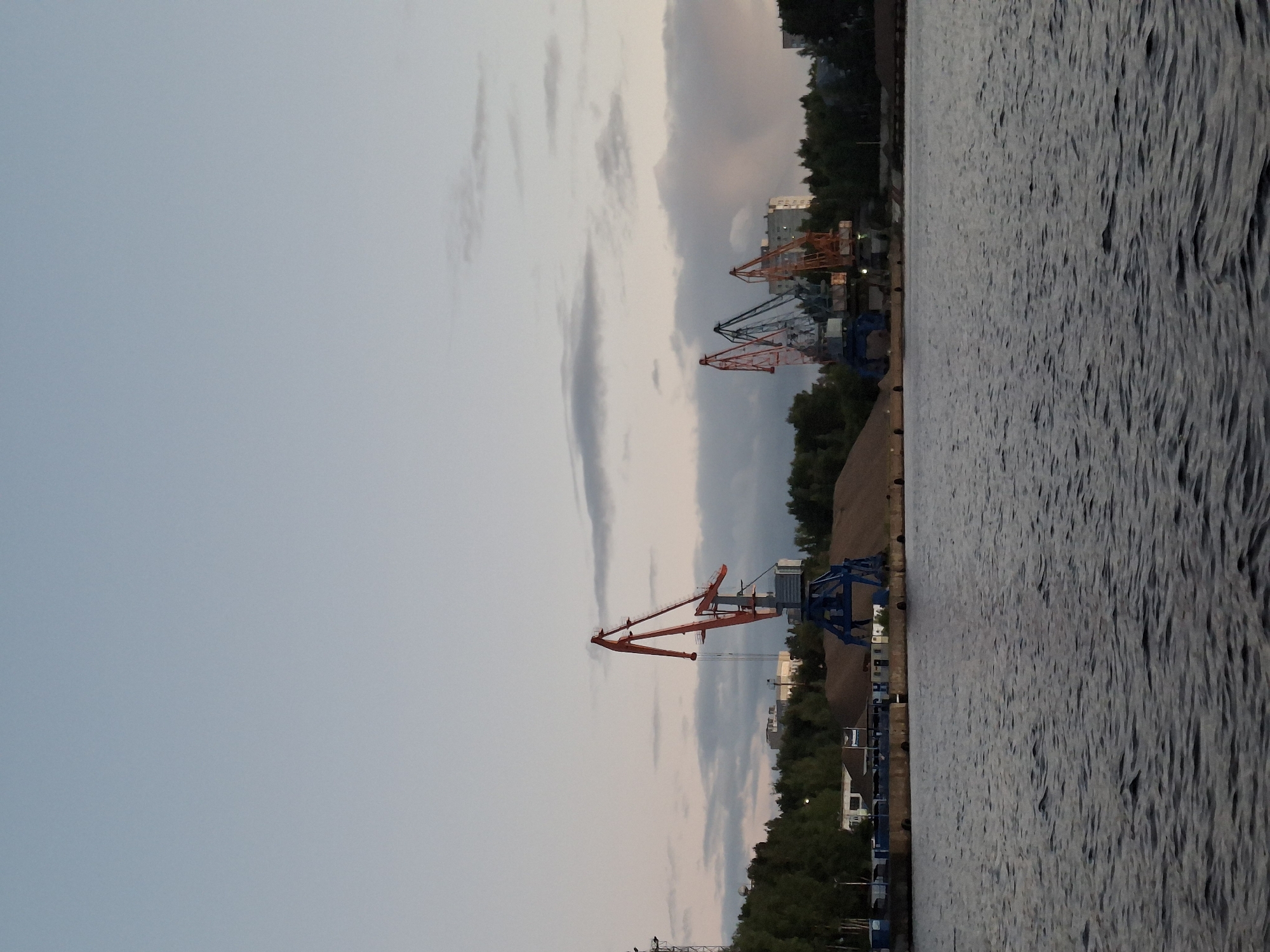 A wide-angle, medium-shot photograph captures a harbor scene at dusk in Chaikovsky, Russia. The sky, a pale blue transitioning to a soft pink near the horizon, is streaked with wispy clouds. Dominating the middle ground are several large port cranes. One prominent red and blue crane stands on the left, with its boom extended. To the right, a cluster of three cranes, two orange and one blue, are visible. Behind the cranes, a large pile of sand or gravel is situated on the embankment. Dense green trees line the background, interspersed with buildings. In the foreground, dark, rippling water fills the bottom half of the frame, reflecting the muted light. There are no people or animals visible in the image. The overall impression is one of industrial activity against a serene twilight sky.