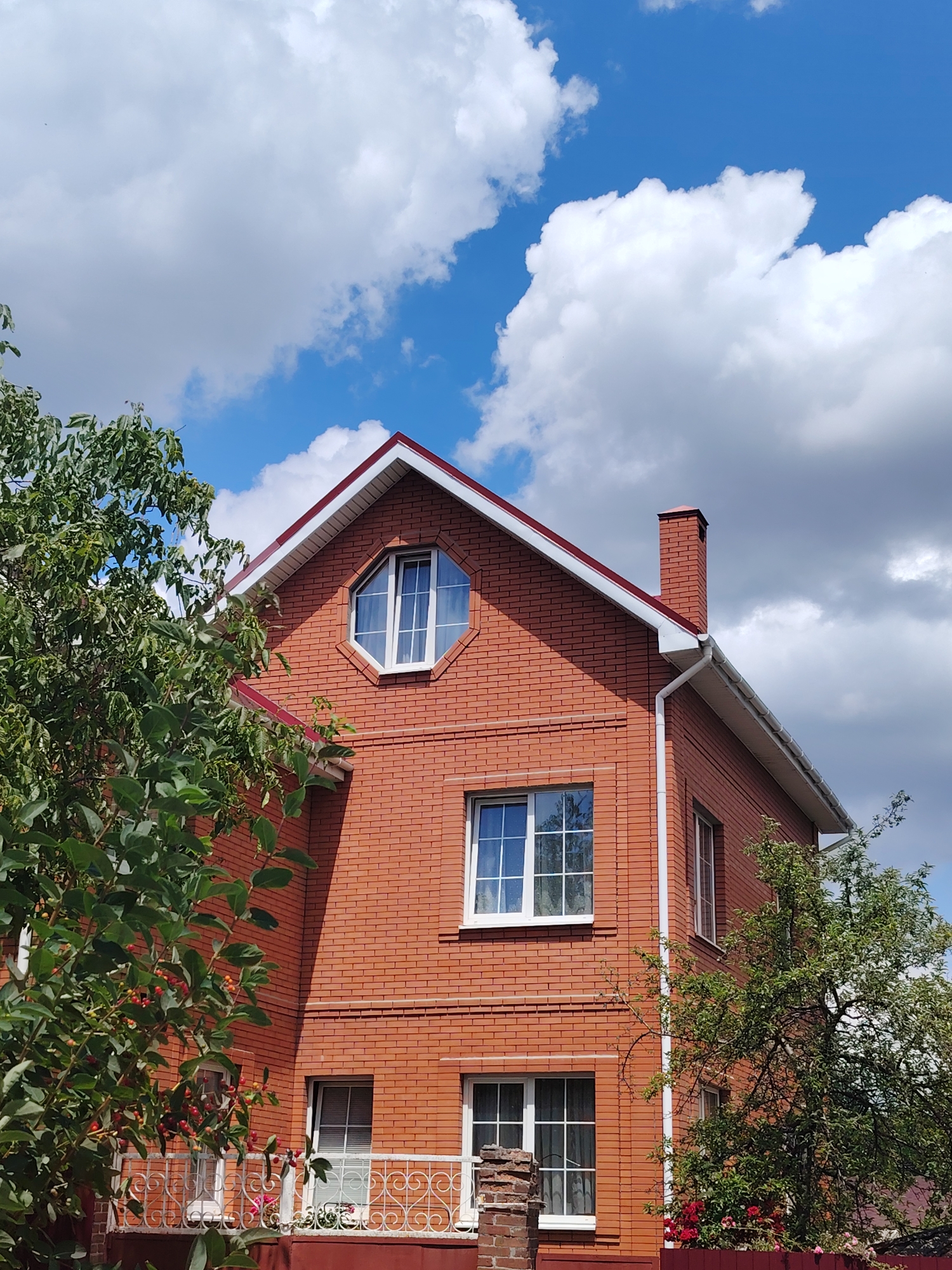 A two-story brick house with a red roof and white trim stands under a blue sky with white, fluffy clouds. The house has several windows, including a distinctive octagonal window on the upper level. A brick chimney rises from the roof. Green trees with some red berries frame the left and right sides of the house. A white metal balcony railing with a heart-shaped pattern is visible in the foreground on the left. The scene appears to be in a residential area, likely in Rostov-on-Don, Russia, given the architectural style and vegetation. There are no people or animals present in the image. The overall impression is one of a sunny, pleasant day in a suburban or rural setting.